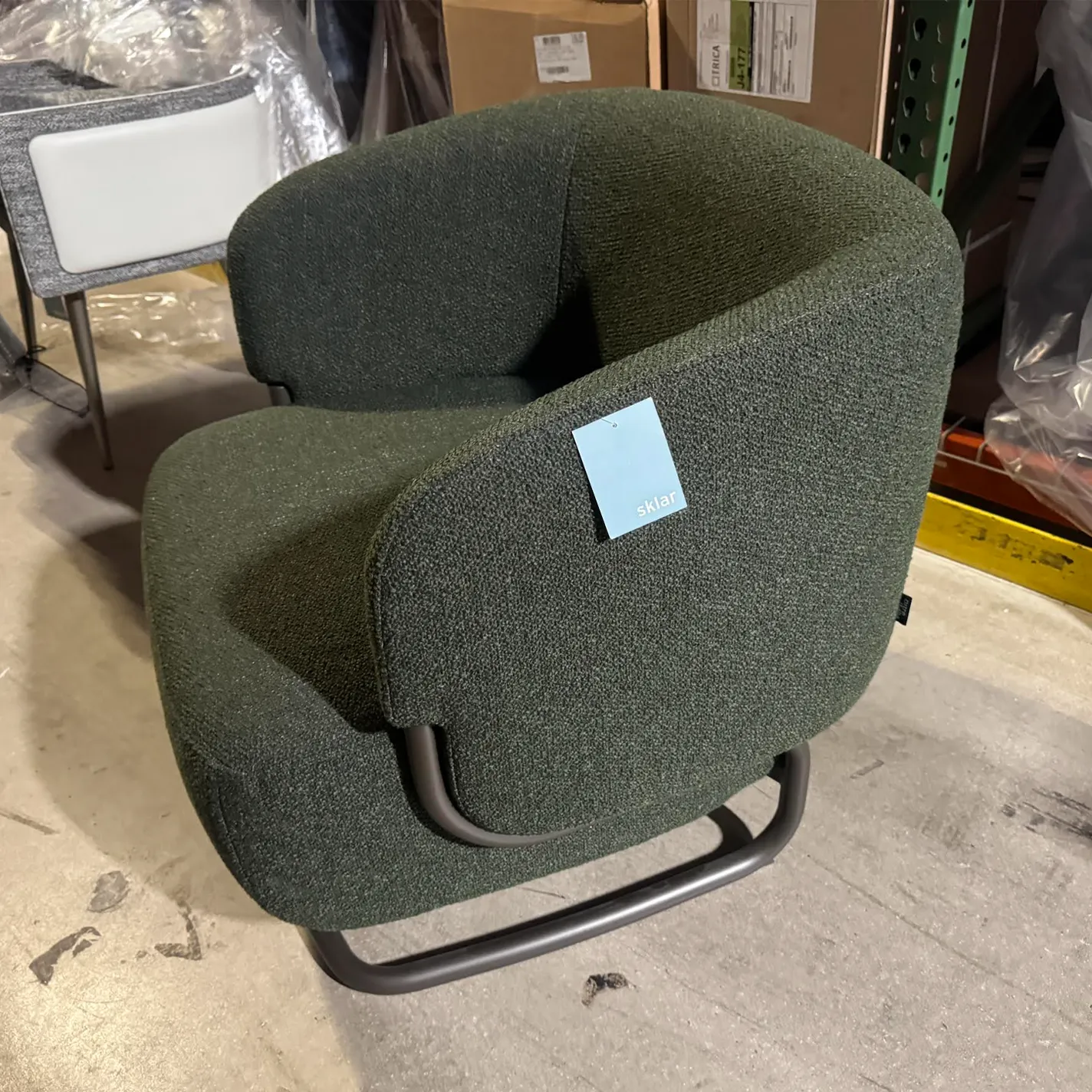 A dark green upholstered armchair with a 'sklar' tag, sitting on a concrete floor in a warehouse.