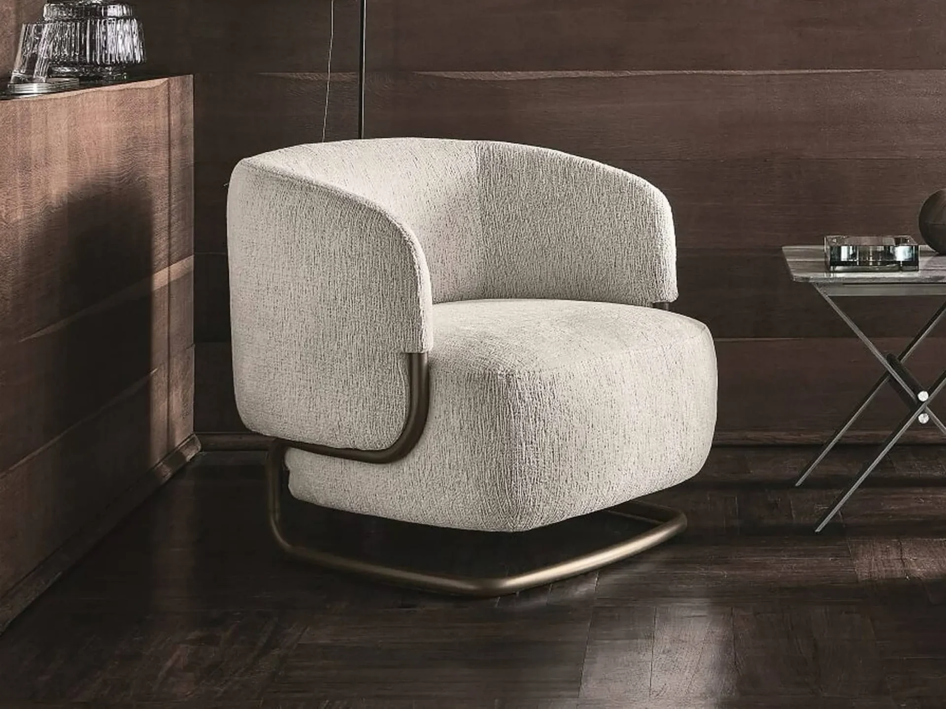 A modern, light beige upholstered armchair with a bronze metal base sits on a dark wooden floor, flanked by a wooden cabinet and a small side table.