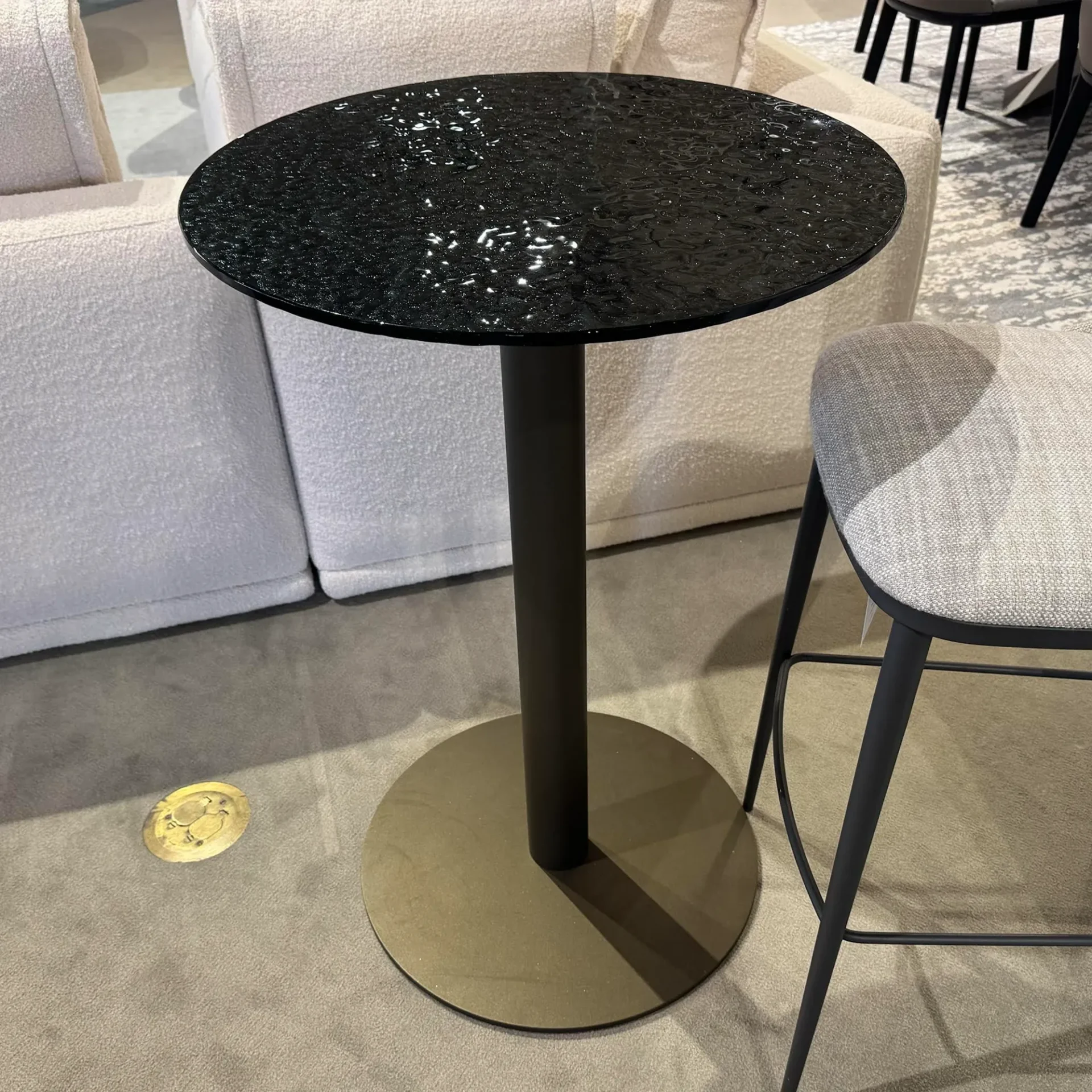 A tall, round table with a textured black top and a dark base, next to a light-colored sofa and a grey bar stool on a grey carpet.
