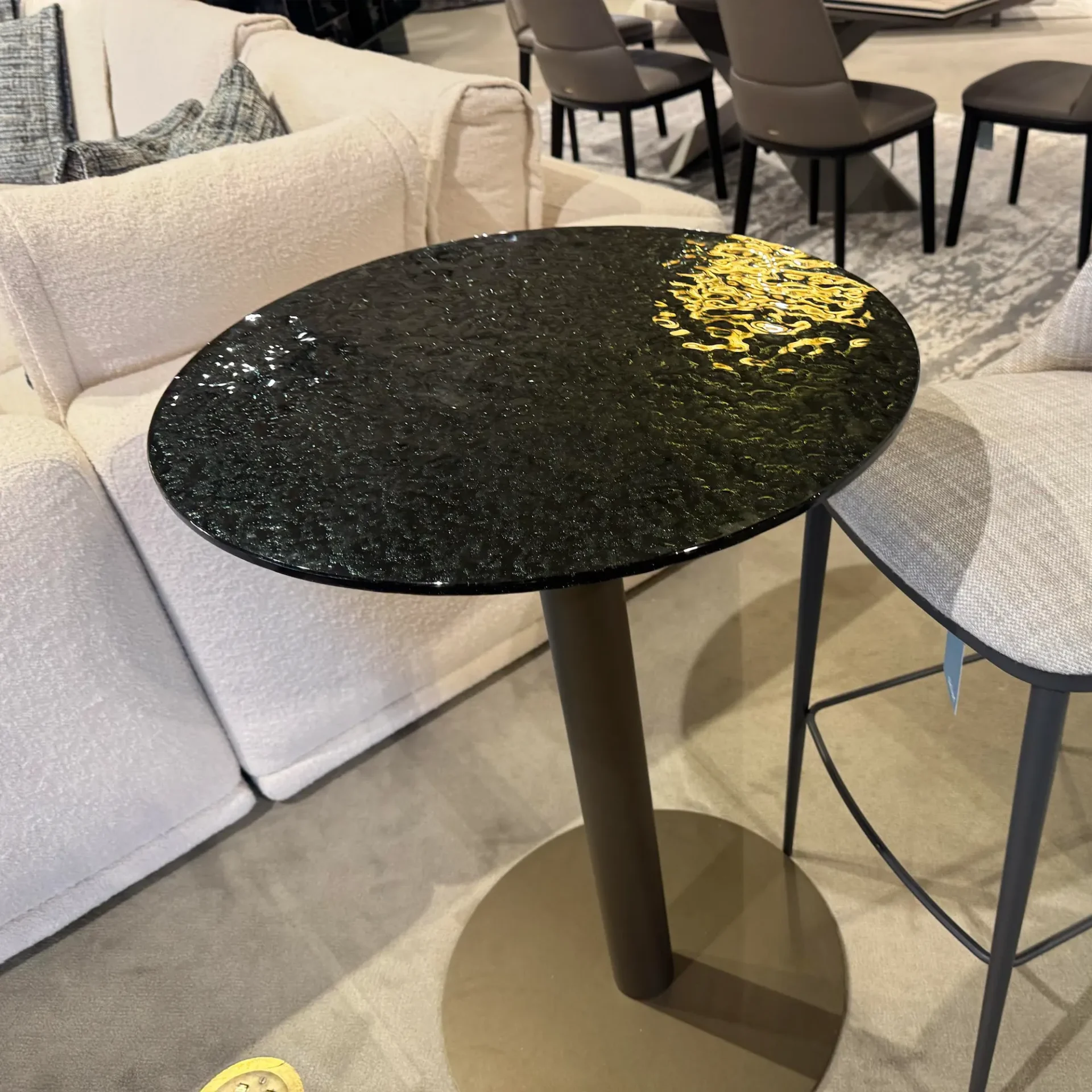 A round, dark, textured side table with a golden reflection, next to a cream sofa and a gray bar stool in a modern room.