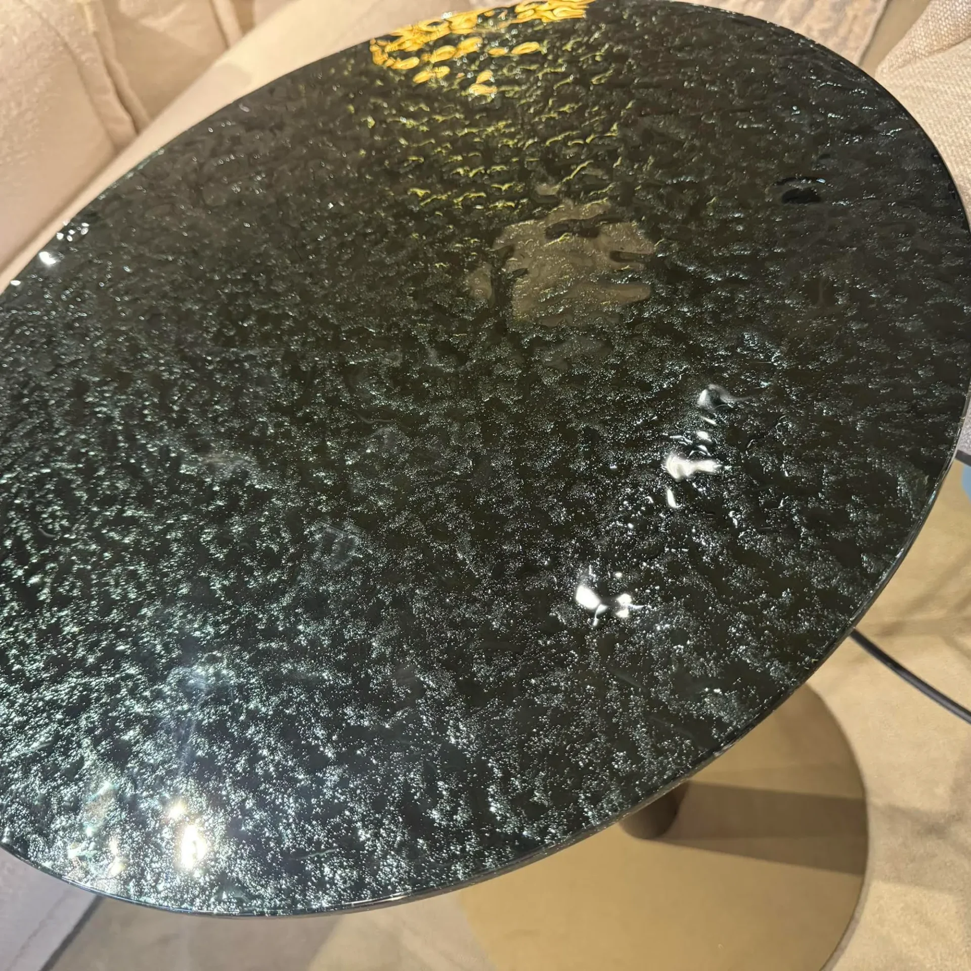A round, dark green or black table with a textured, reflective surface, showing light reflections and a partial view of a light-colored sofa in the background.