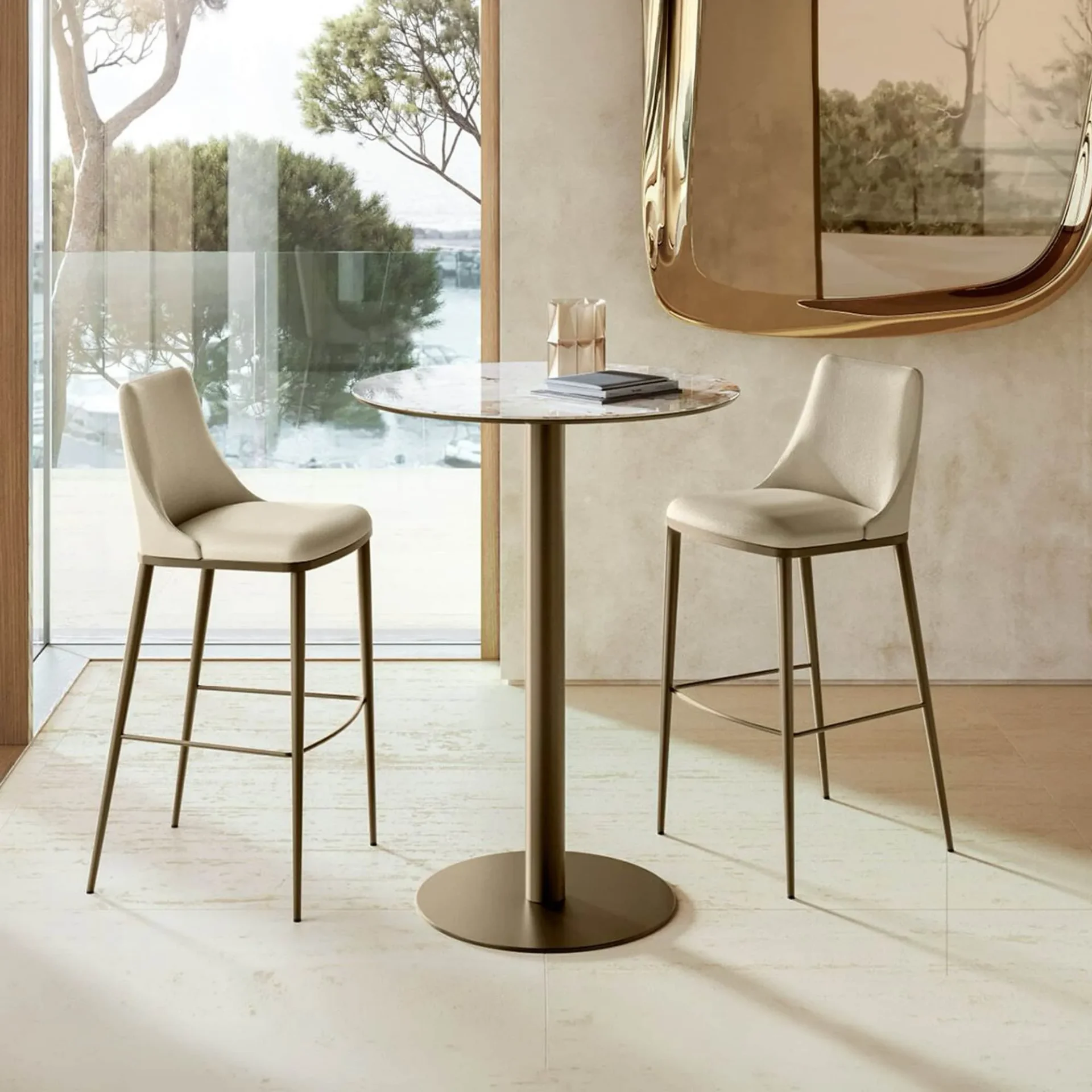 A modern bar table with a marble top and two beige upholstered bar stools in front of a large window overlooking trees and water.