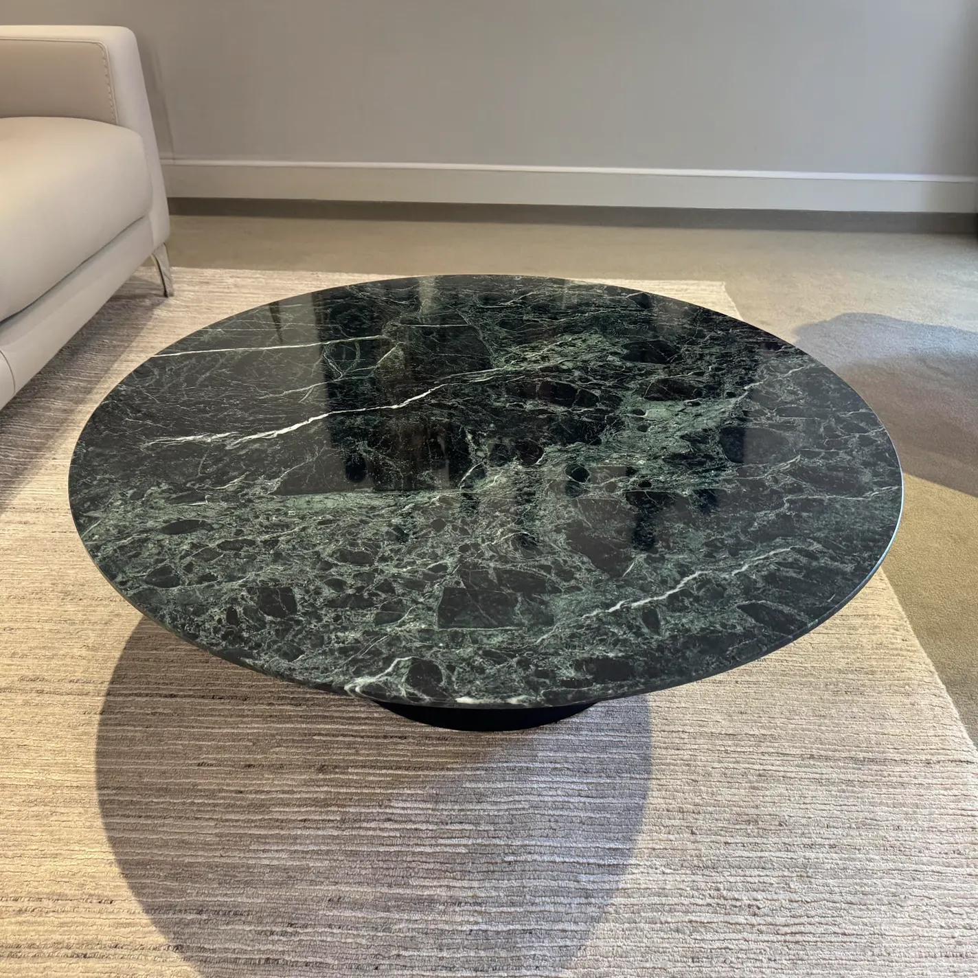 A round dark green marble coffee table with white veins sits on a light textured rug, next to a cream sofa.