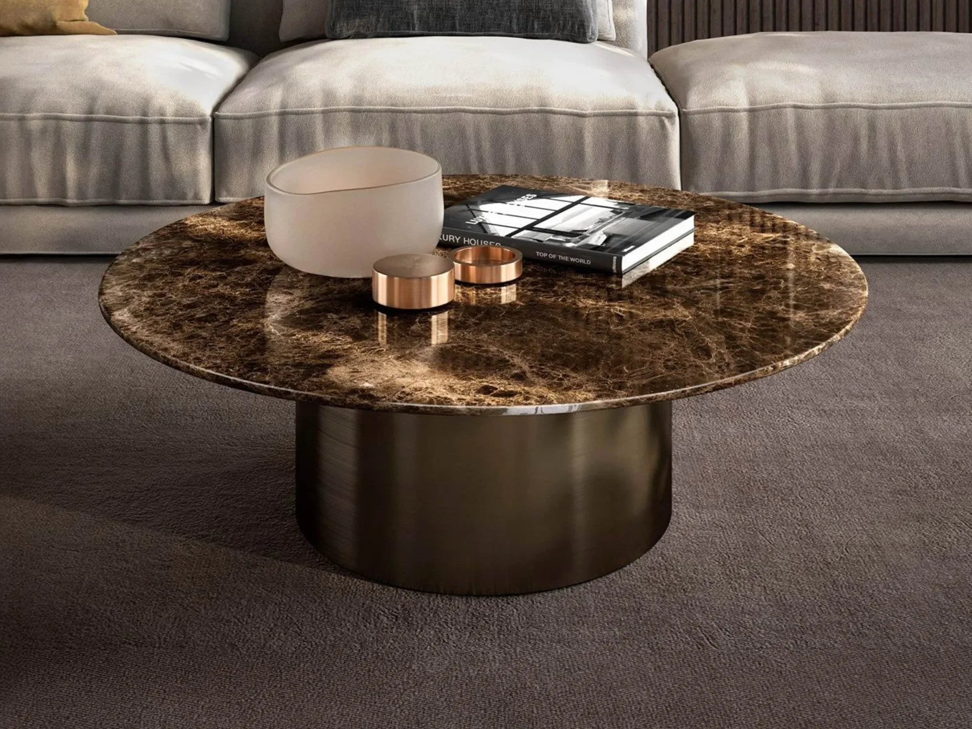 A round brown marble coffee table with a dark bronze base, holding a frosted vase, two copper-colored objects, and a black book, in front of a light gray sofa.