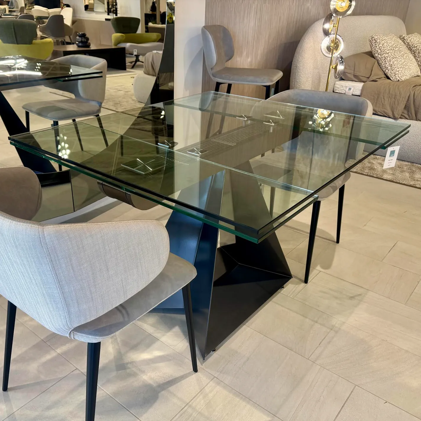 A modern rectangular glass dining table with a dark geometric base, surrounded by light gray upholstered chairs, set in a contemporary furniture showroom.