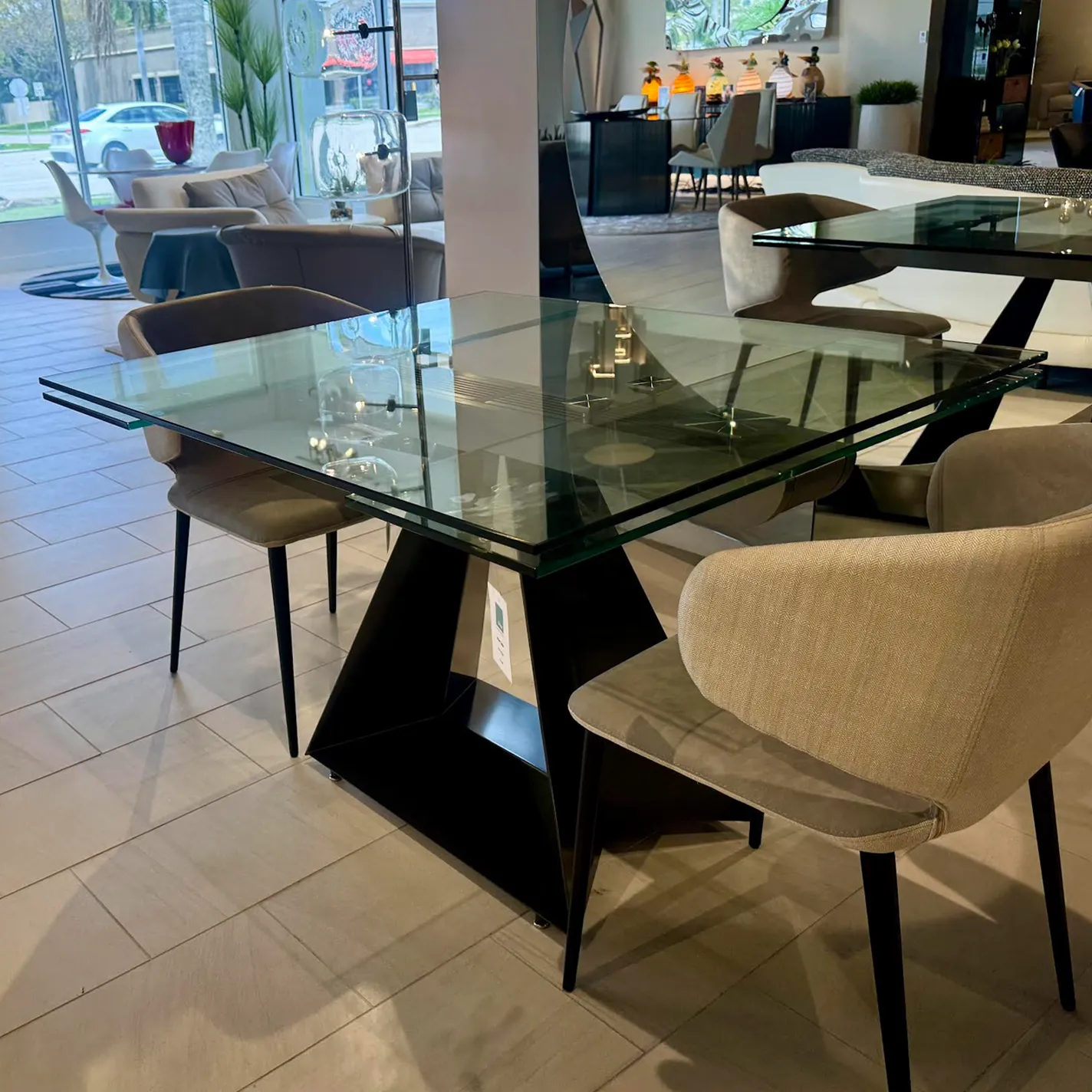A modern square glass dining table with a black geometric base and four beige upholstered chairs in a furniture showroom.