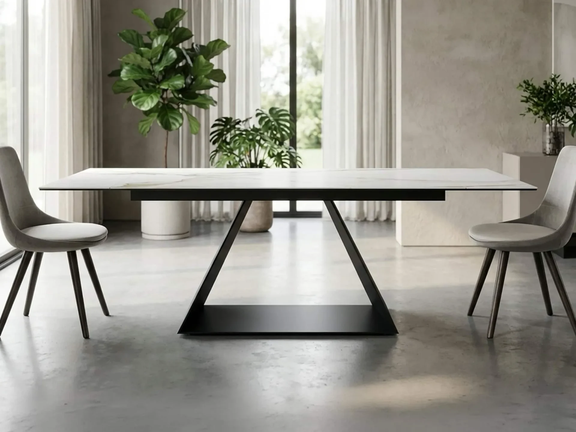 A modern dining table with a white marble top and black geometric base, flanked by two grey upholstered chairs, in a bright room with large windows and potted plants.