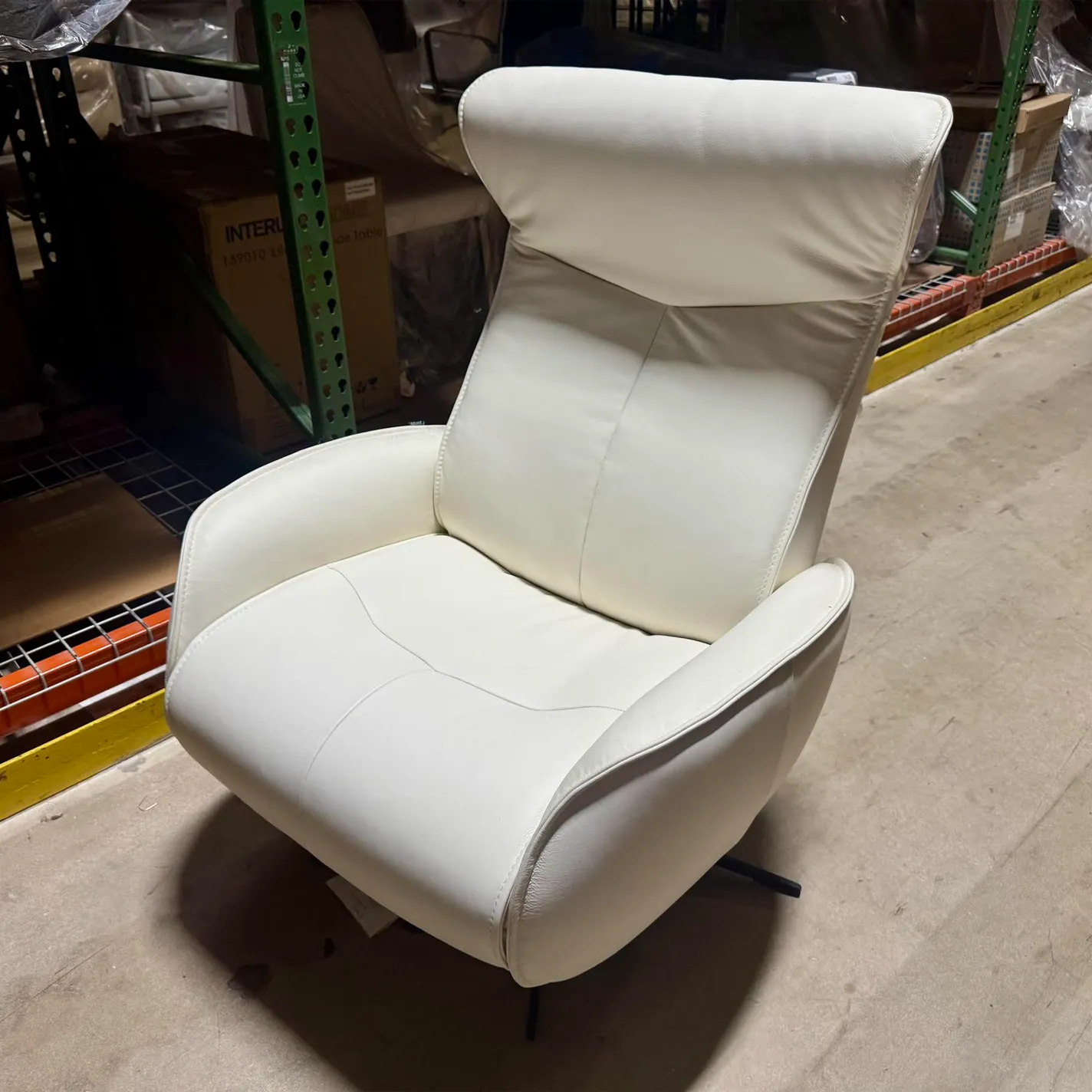 A cream-colored modern recliner chair with a high back and padded armrests, set against a warehouse background with shelving and boxes.