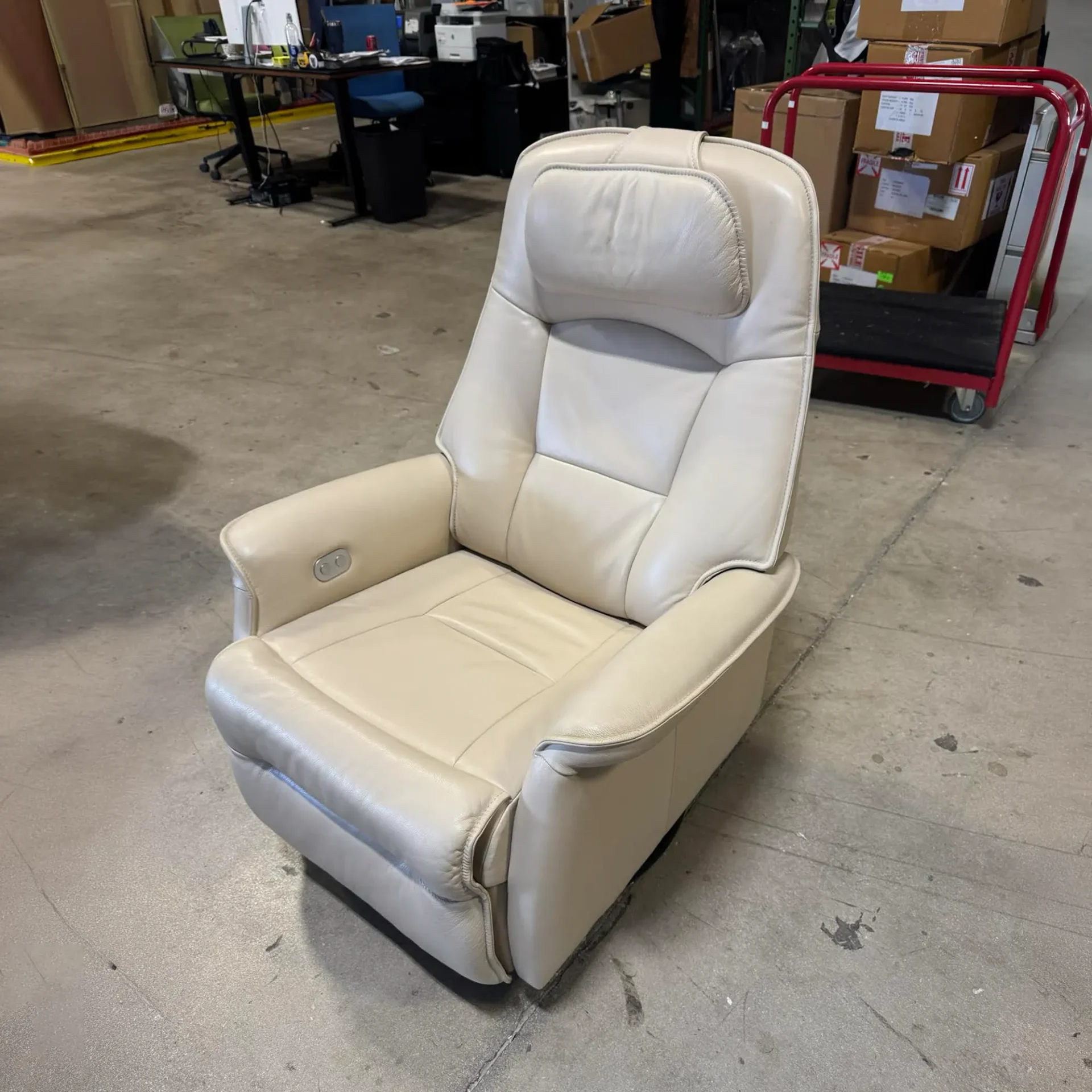 A cream leather recliner chair on a concrete floor, with office equipment and stacked boxes in the background.