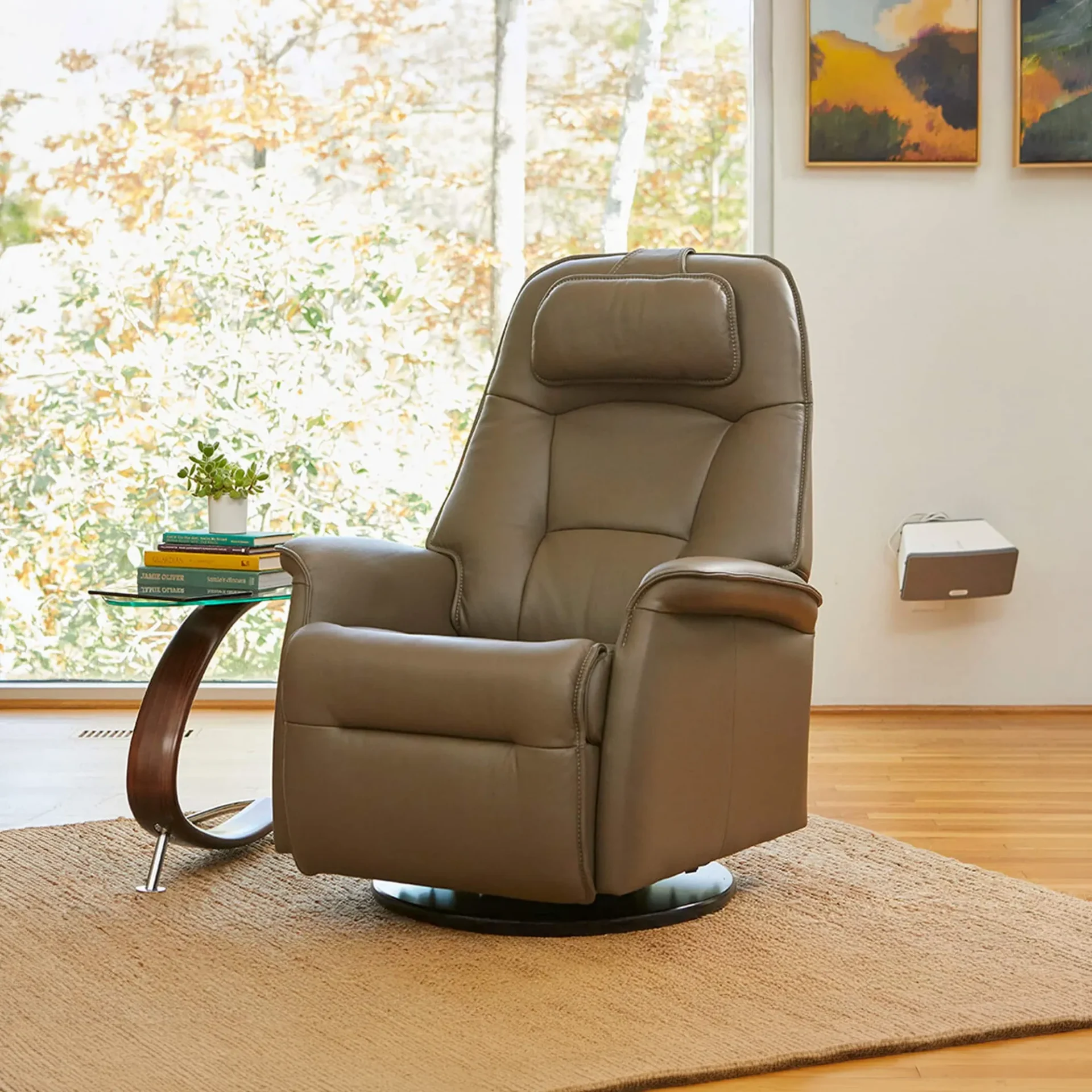 A taupe leather recliner chair on a rug, next to a glass-top table with books and a plant, with a window view of trees and artwork on the wall.