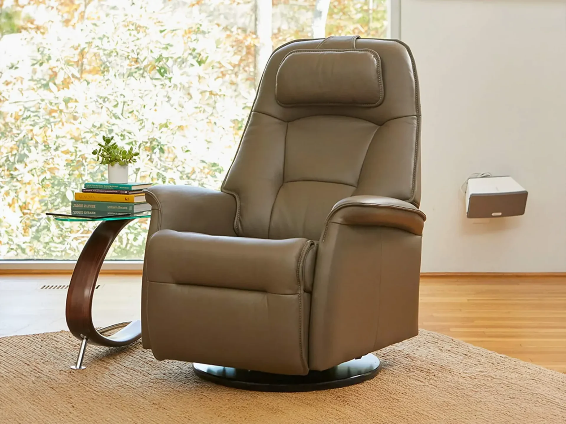A taupe leather recliner chair on a rug, next to a glass side table with books and a plant, in a brightly lit room with a window.