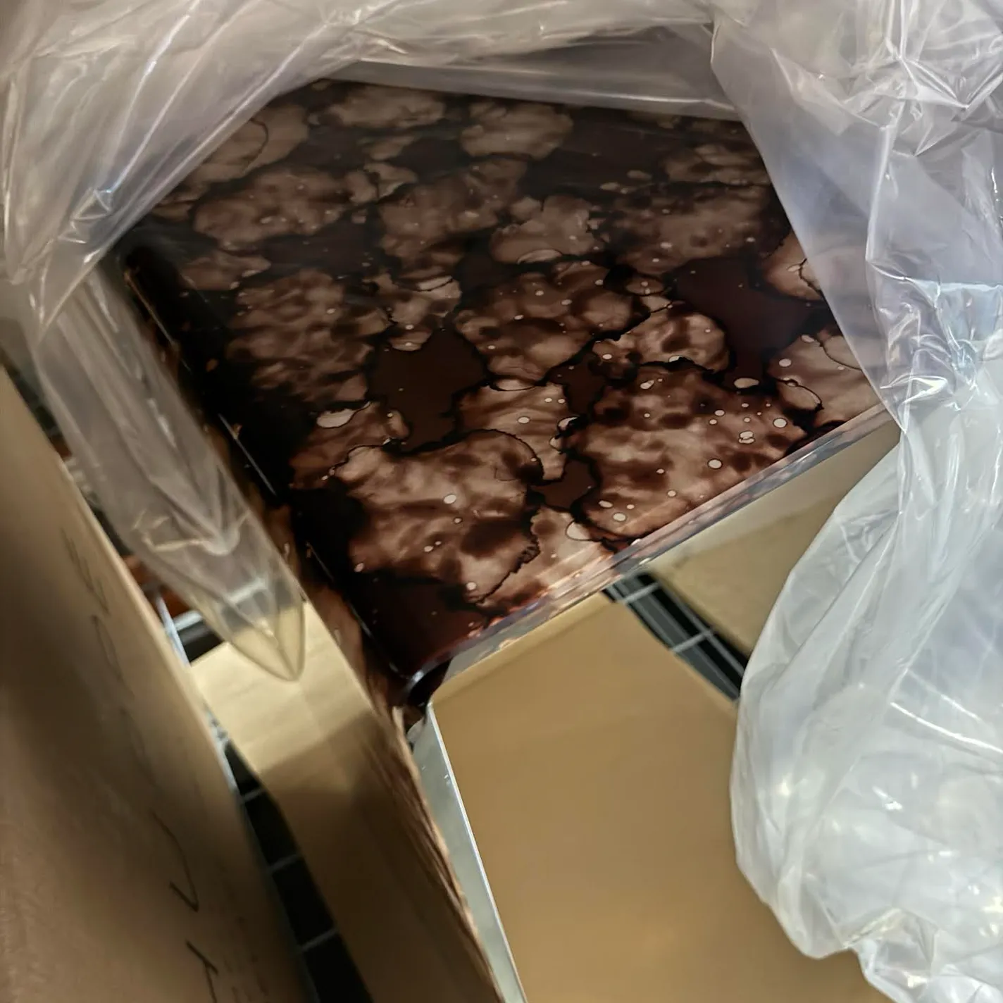 A dark brown surface with light marbled patterns and white speckles, partially covered in clear plastic wrap, inside a cardboard box.