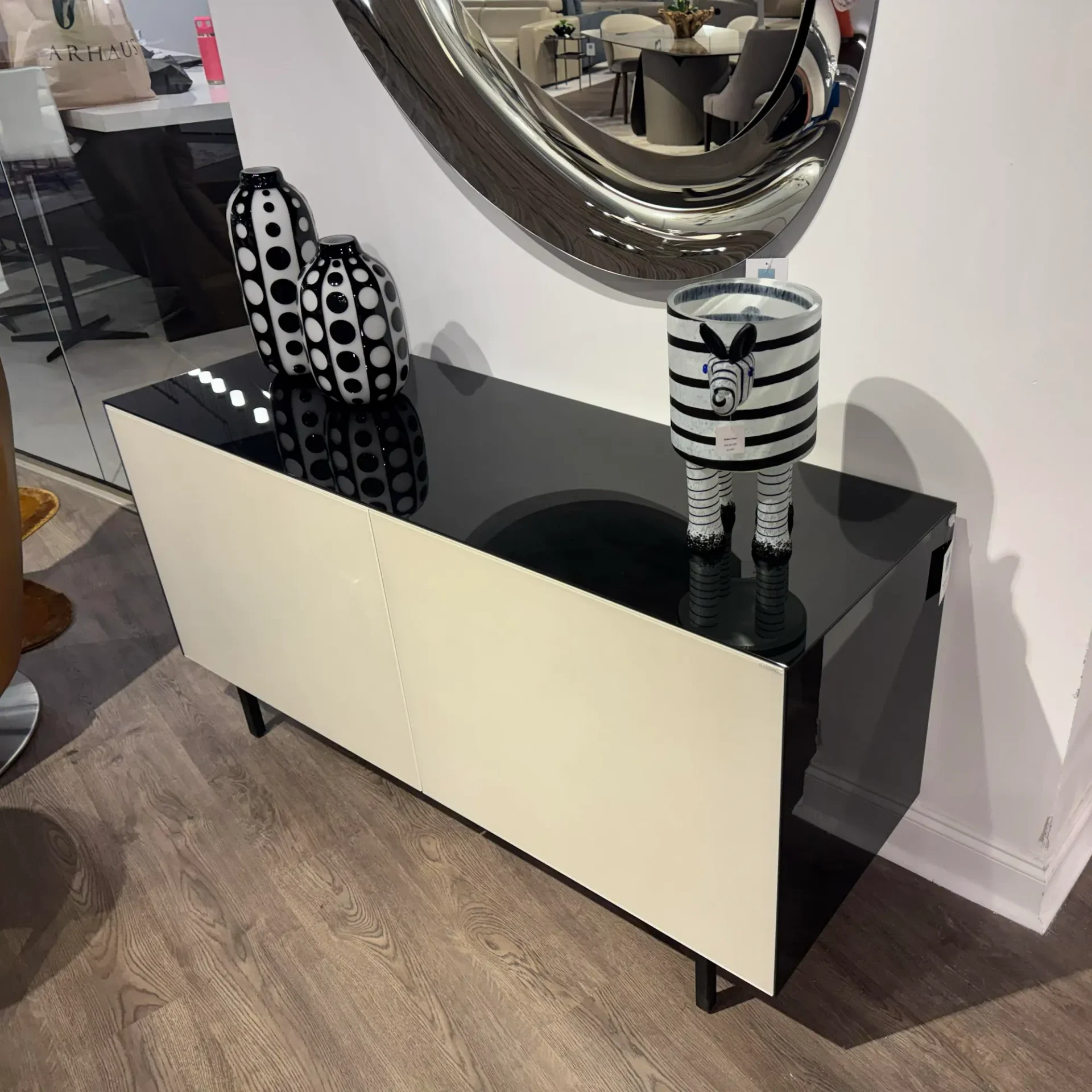 A modern black and cream sideboard with a glossy top, adorned with black and white polka-dotted vases and a striped zebra figure, beneath a large metallic mirror.