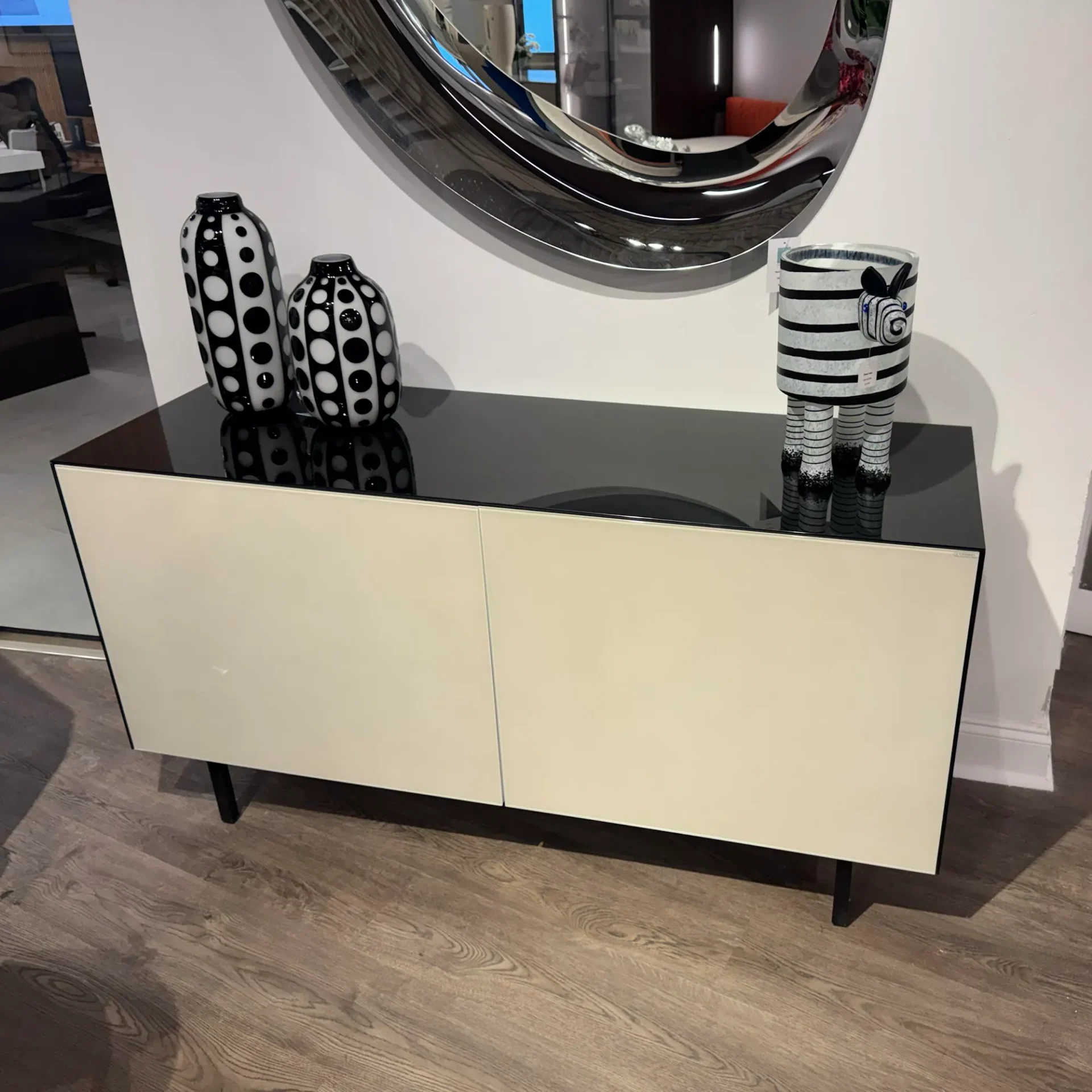 A cream and black modern sideboard with two polka-dotted vases and a zebra sculpture on top, beneath a wavy mirror.