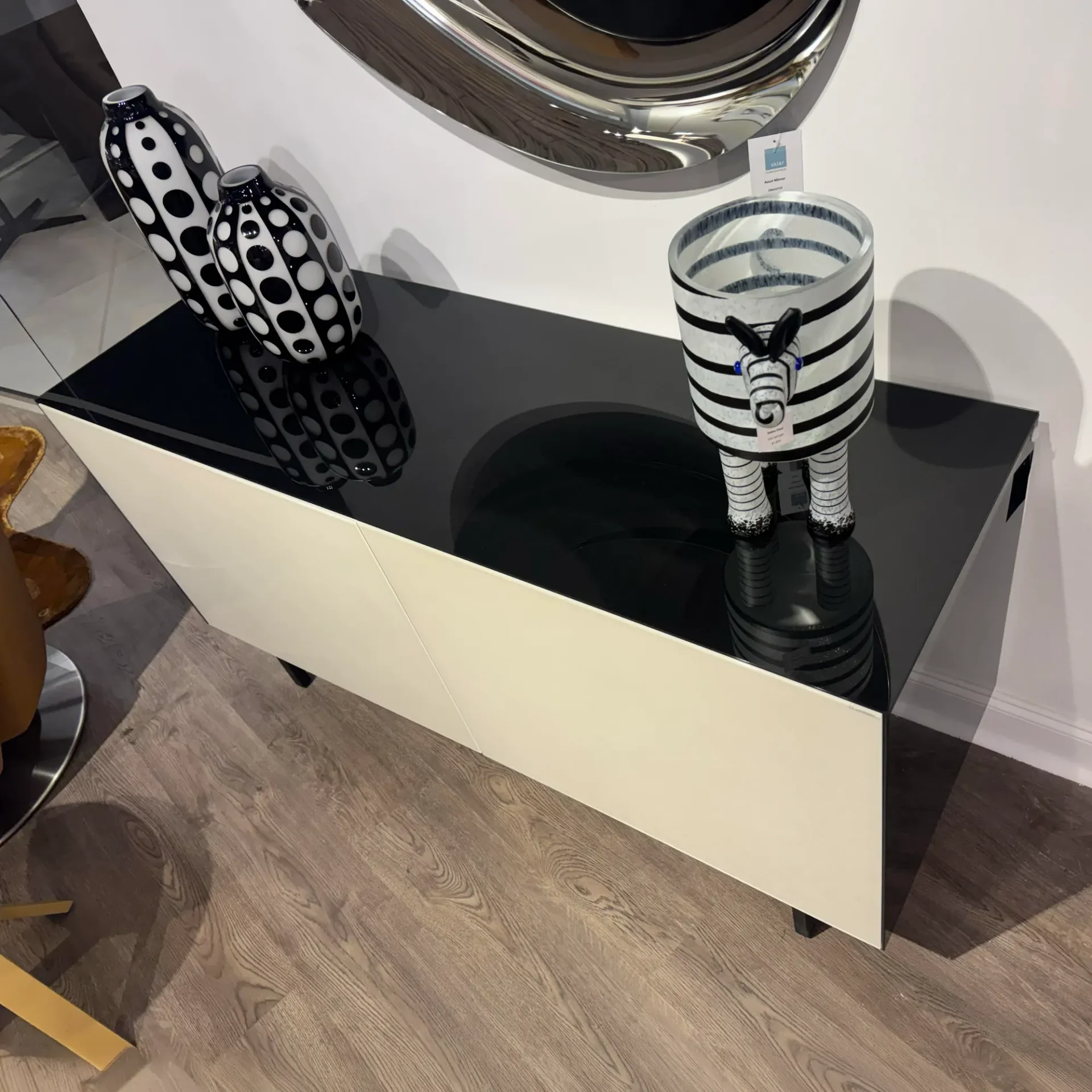 A modern console table with a black glass top and off-white body, displaying two black and white polka-dotted vases and a zebra-striped decorative cylinder. A large silver mirror hangs above.