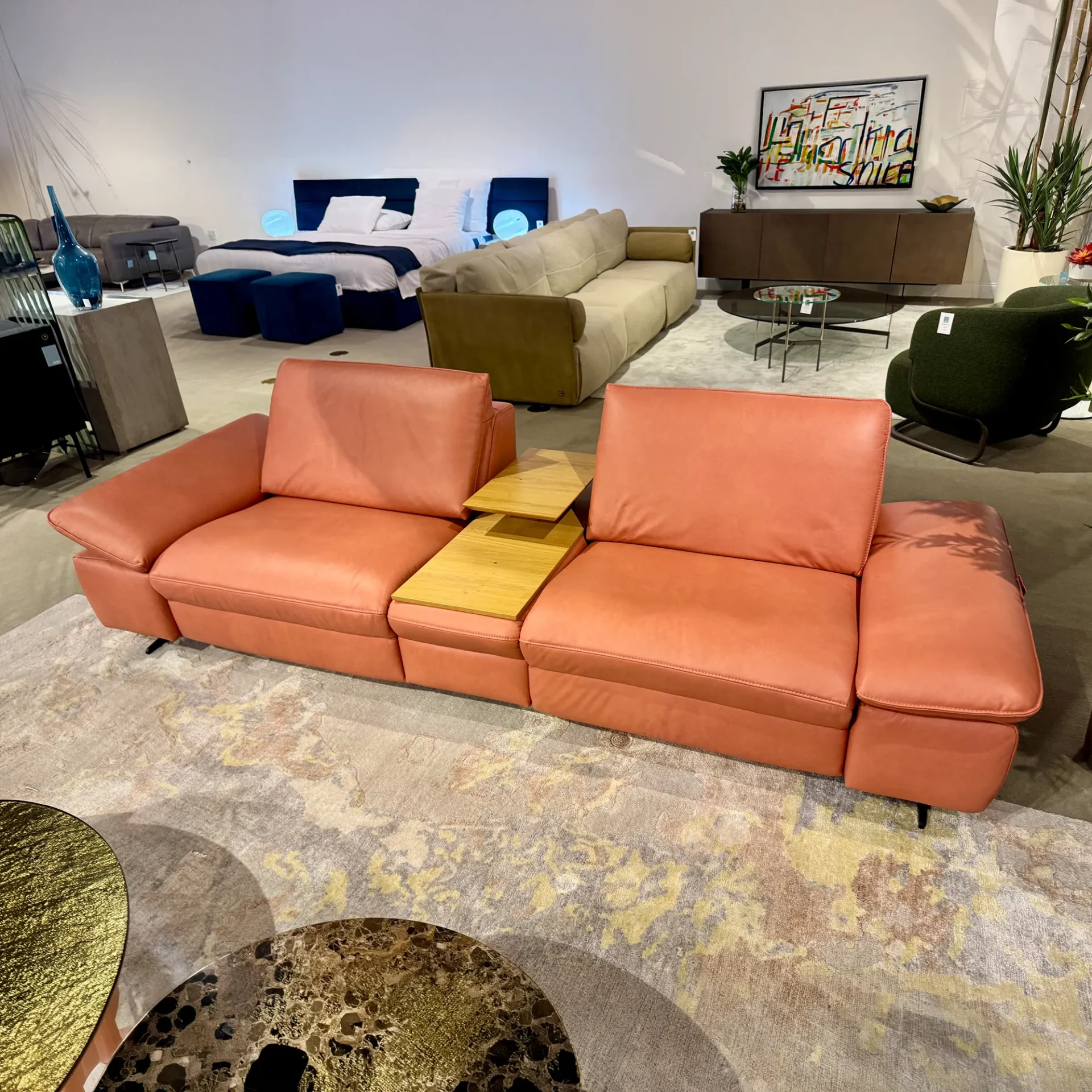 A coral modular sofa with integrated wooden tables on a patterned rug in a modern furniture showroom, with other furniture arrangements in the background.