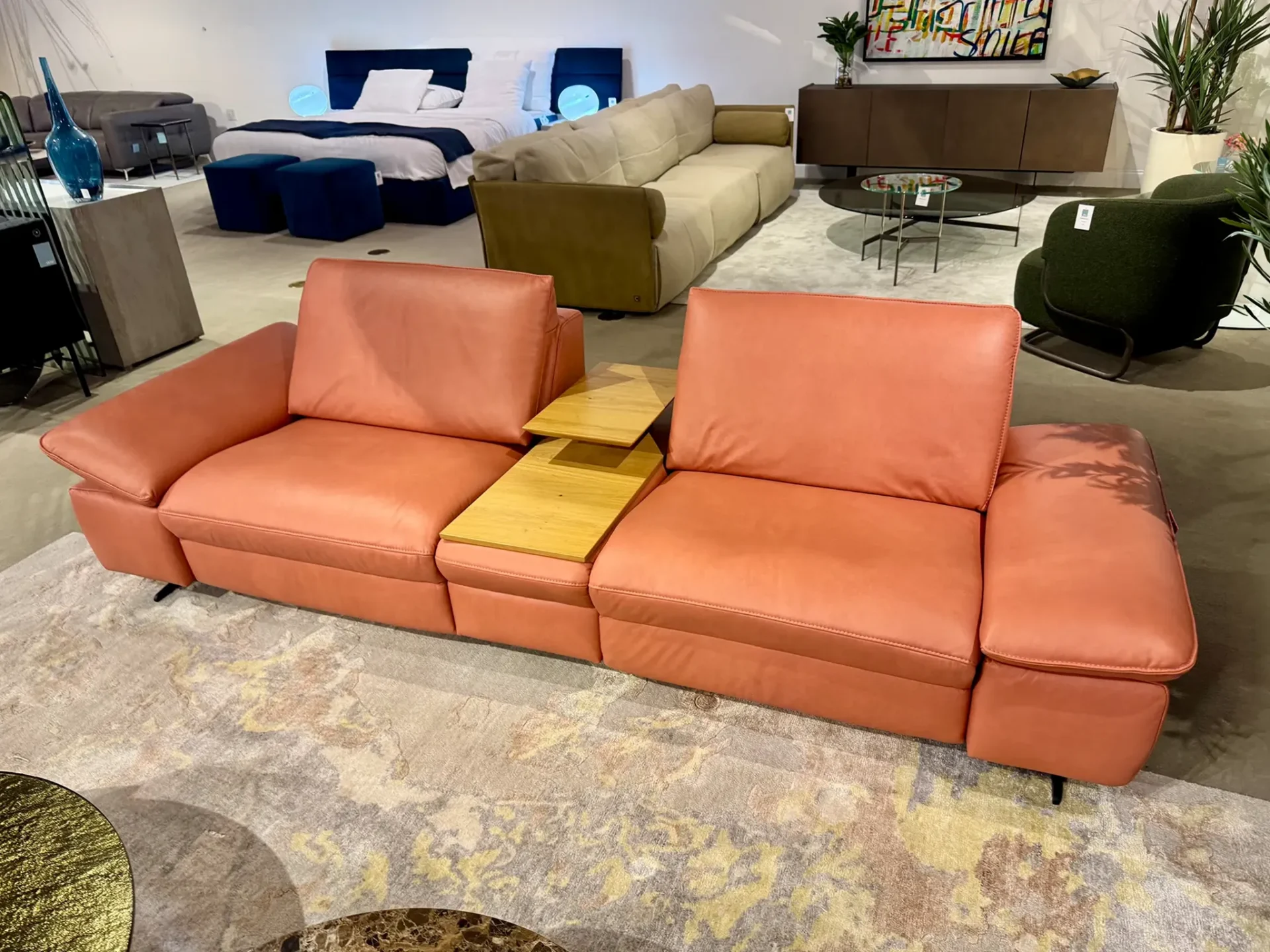 A large, modern coral-colored leather sofa with a wooden tray table in a furniture showroom, surrounded by other beds, sofas, and decor.
