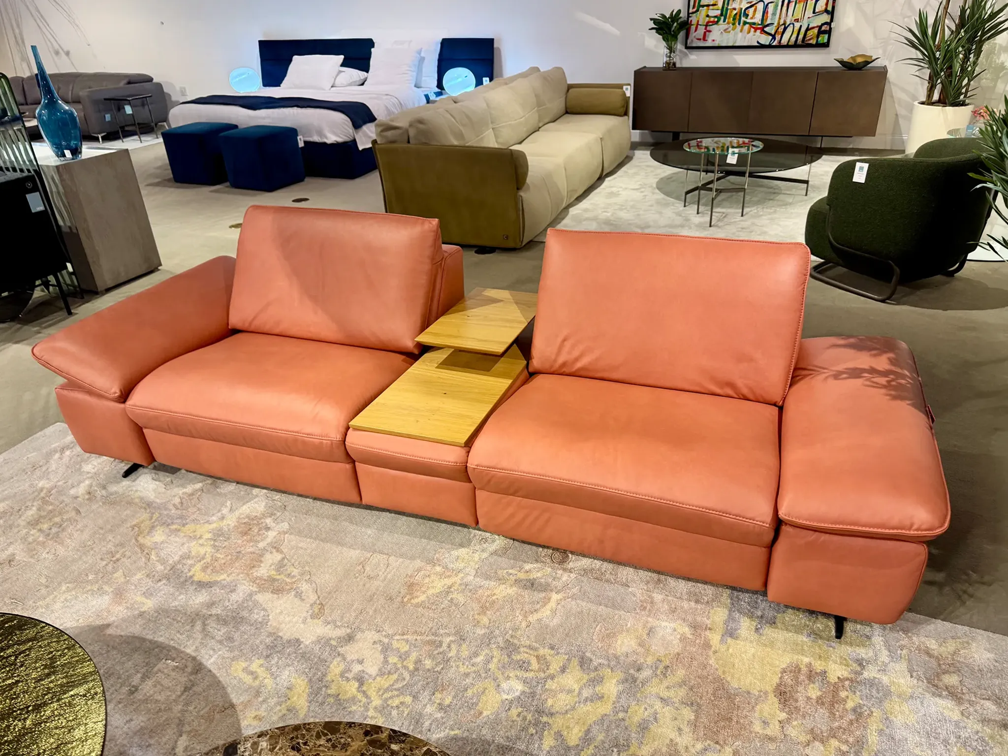 A large, modern coral-colored leather sofa with a wooden tray table in a furniture showroom, surrounded by other beds, sofas, and decor.