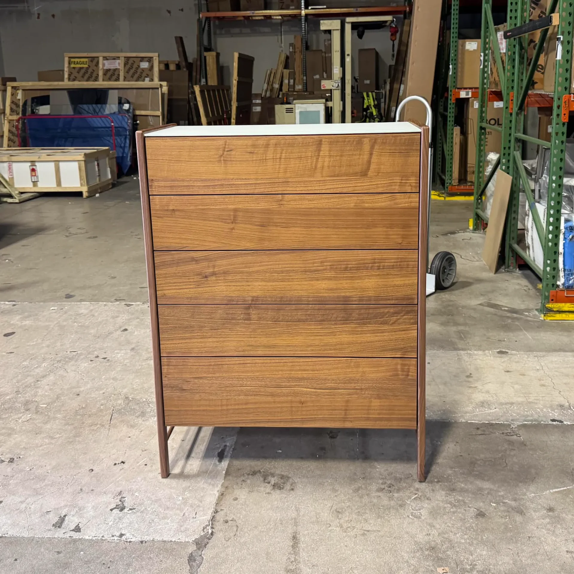 A five-drawer wooden dresser with a white top and brown frame stands on a concrete floor in a warehouse setting.