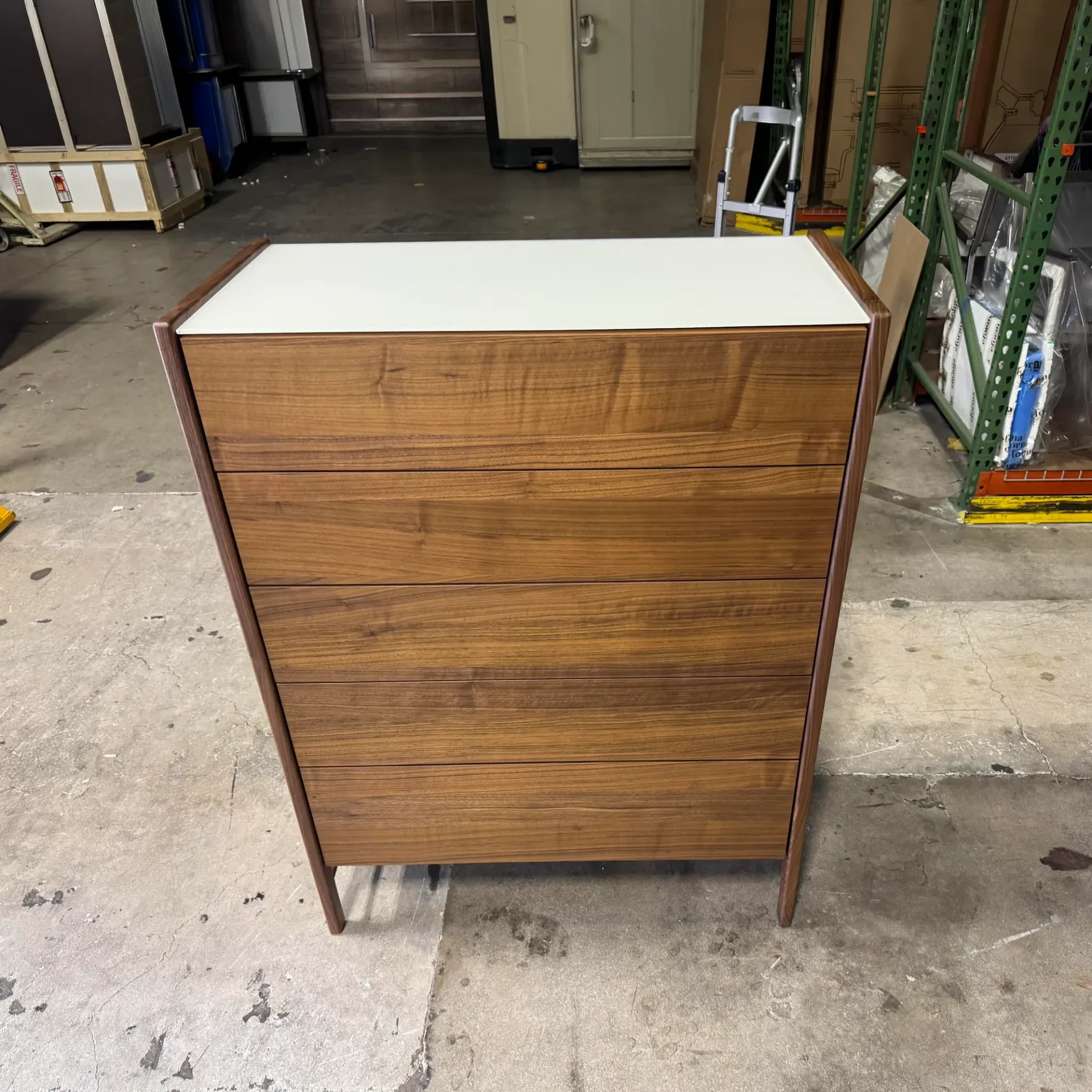 A tall, narrow modern chest of drawers with a white top and five warm wood grain drawers, framed by darker wood, stands on a concrete floor in a warehouse.