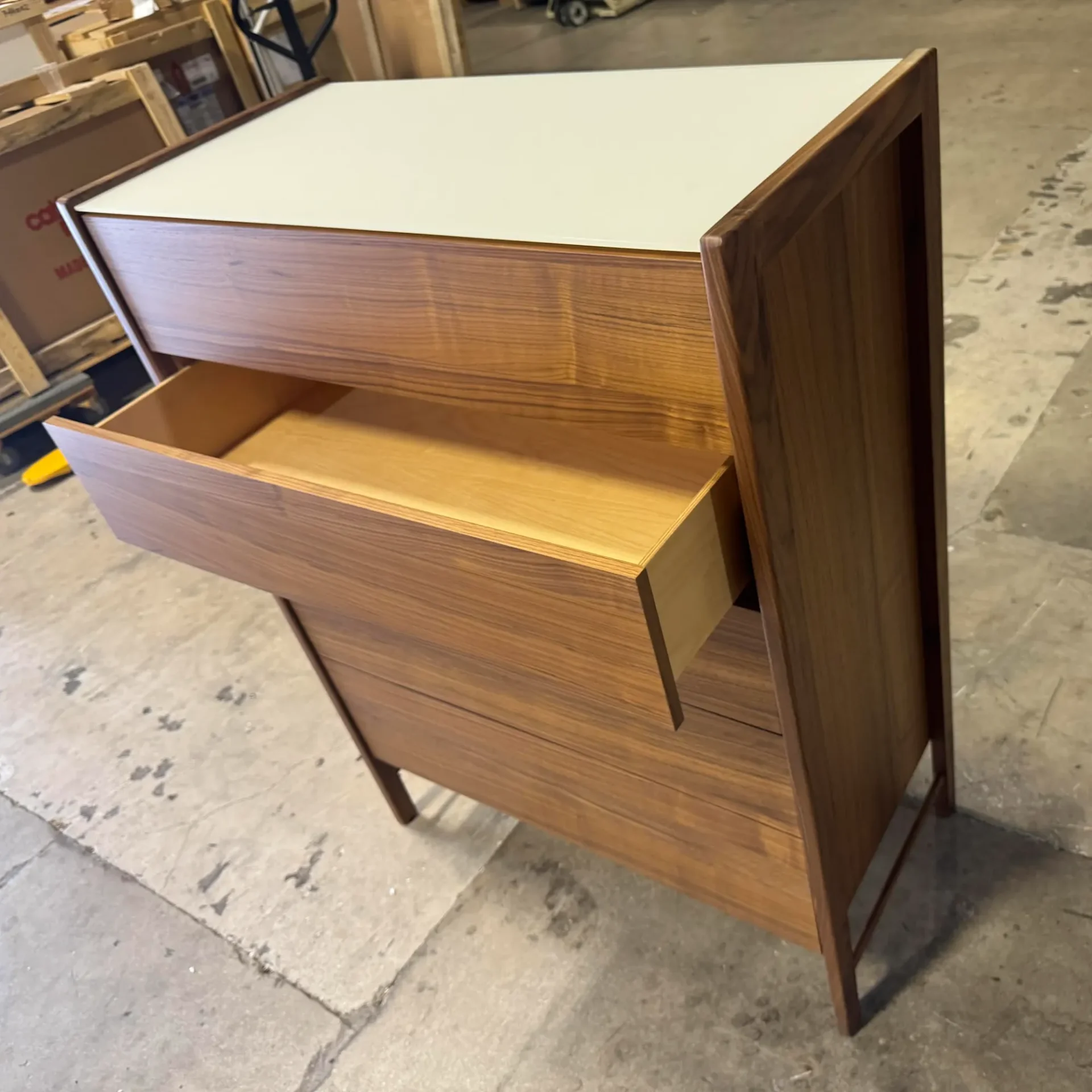 A wooden dresser with a light top and four drawers, with the top drawer open, on a concrete floor in a warehouse.