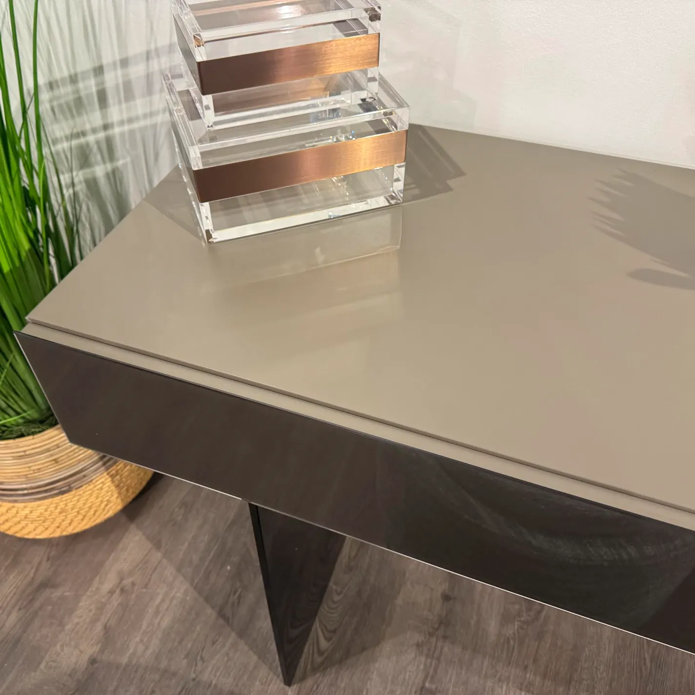 A modern console table with a glossy taupe top, dark front panel, green plant in a basket, and stacked clear acrylic boxes with copper bands.