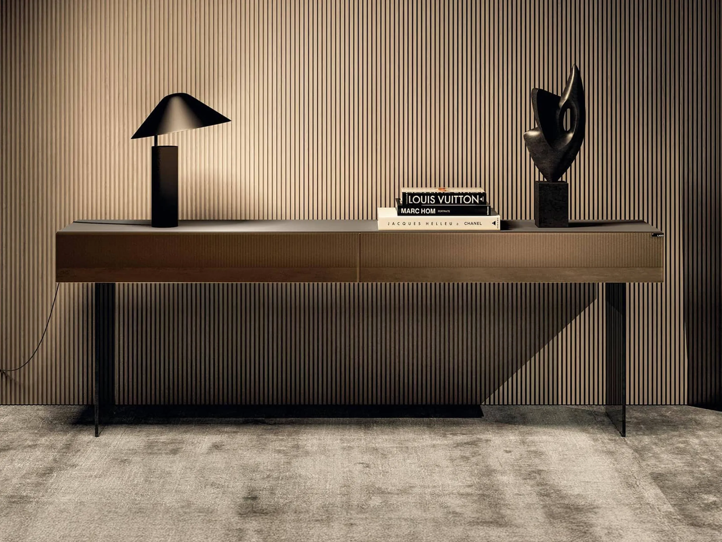 A modern console table with a black lamp, a stack of fashion books, and an abstract dark sculpture, set against a ribbed wooden wall and a textured gray rug.