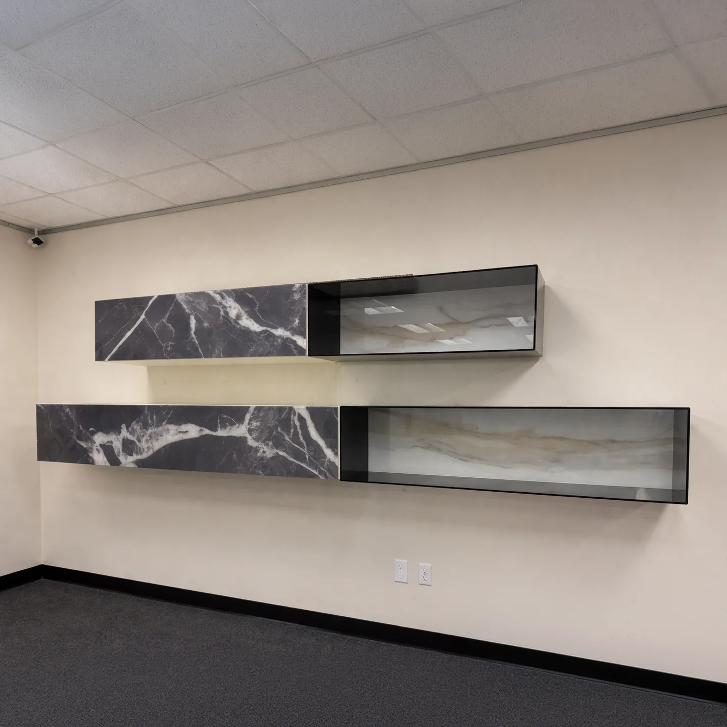 Two sets of modern wall-mounted shelves against a light wall. Each set has a dark grey marble-patterned cabinet next to a black-framed open shelf with a translucent, wavy light beige and white pattern. Dark grey carpet and a white acoustic tile ceiling are visible.