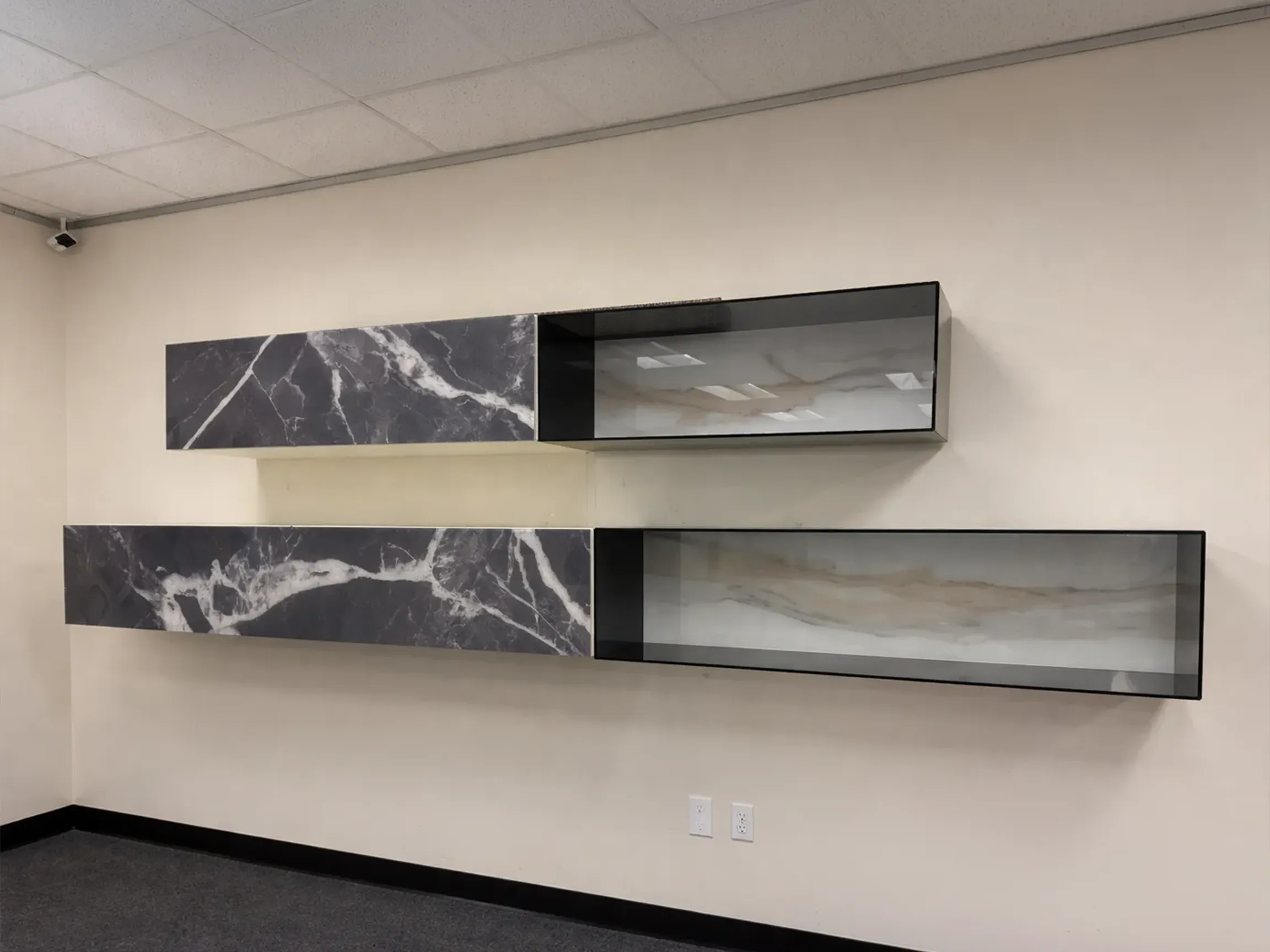 Two rows of wall-mounted units. Each row has a dark gray marble-patterned cabinet and an open shelf with a black frame and light marble back panel. The wall is light beige, the floor is dark gray carpet, and a security camera is in the top left.