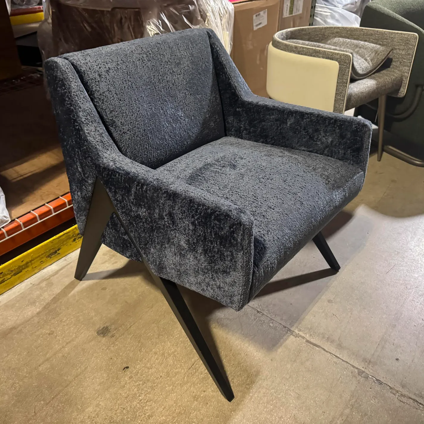 A dark gray upholstered armchair with splayed dark legs sits on a concrete floor in a warehouse setting. Other furniture and boxes are in the background.