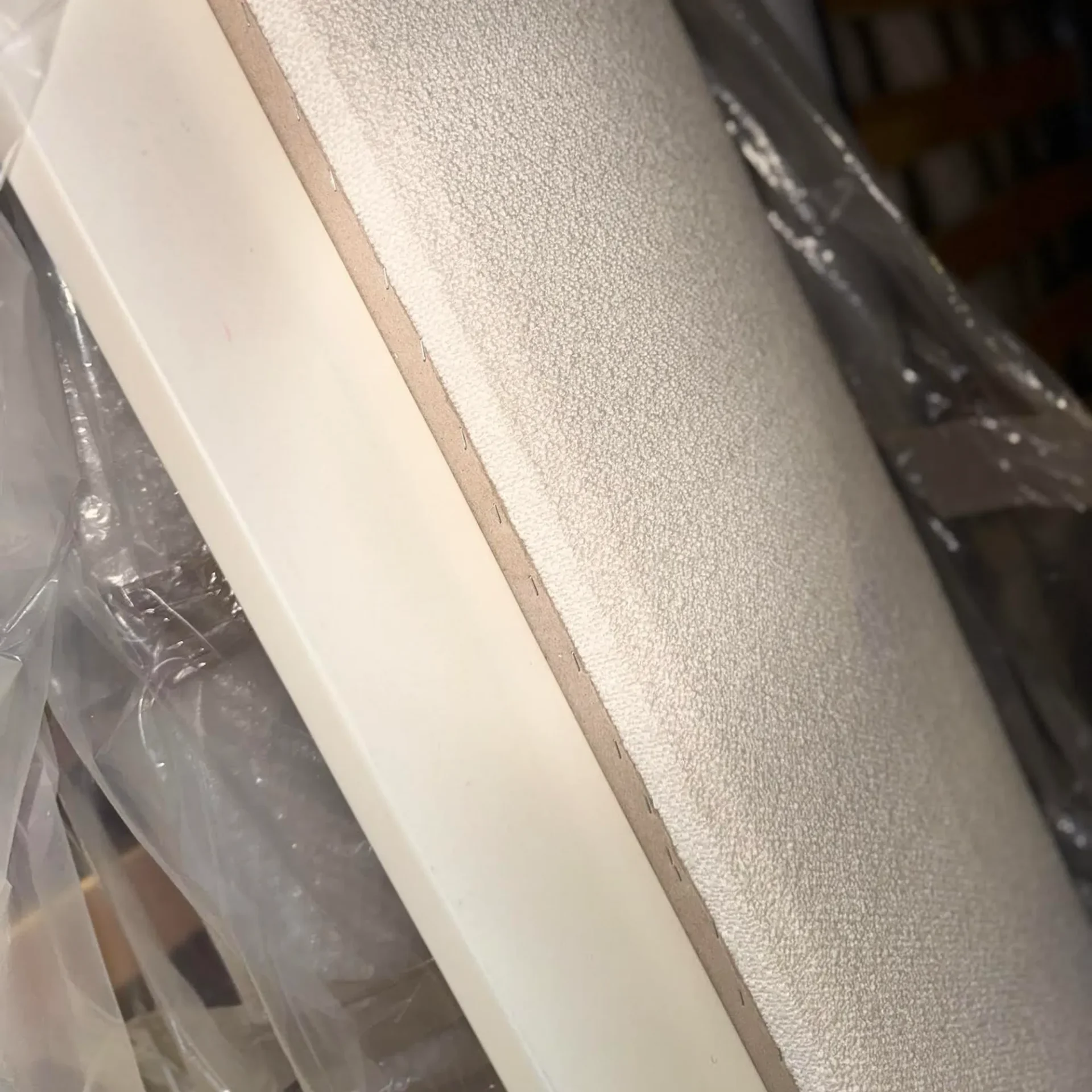 Close-up of a panel with a textured light gray surface and a smooth off-white edge, secured with staples and partially covered in clear plastic.