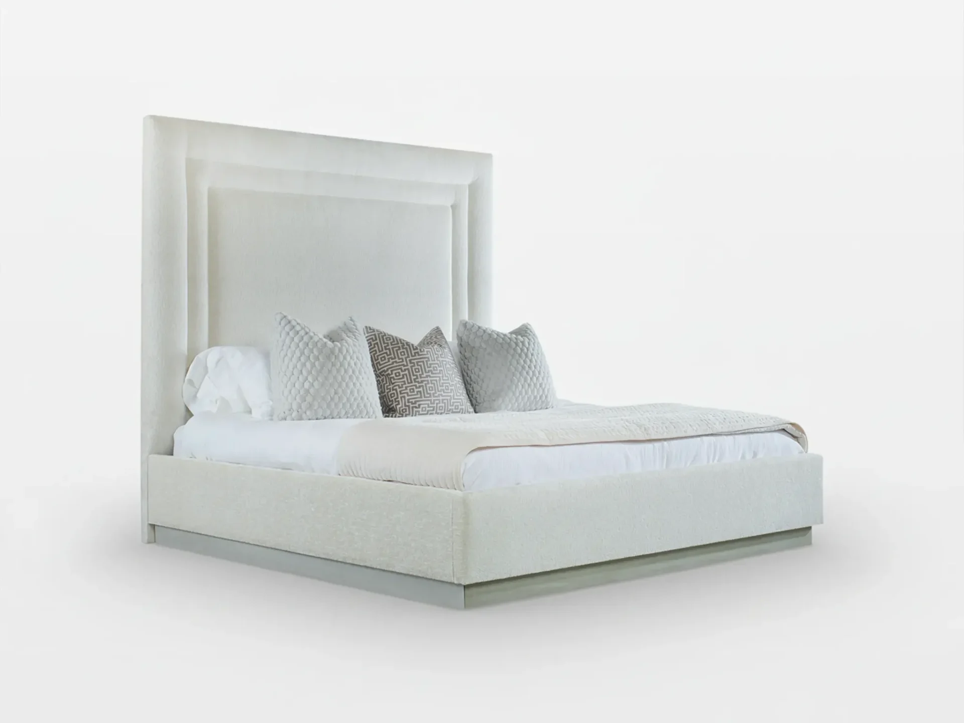 A modern upholstered bed with a tall, layered headboard in a light, textured fabric. The bed is dressed with white sheets, three decorative pillows, and a throw blanket, all in neutral tones.