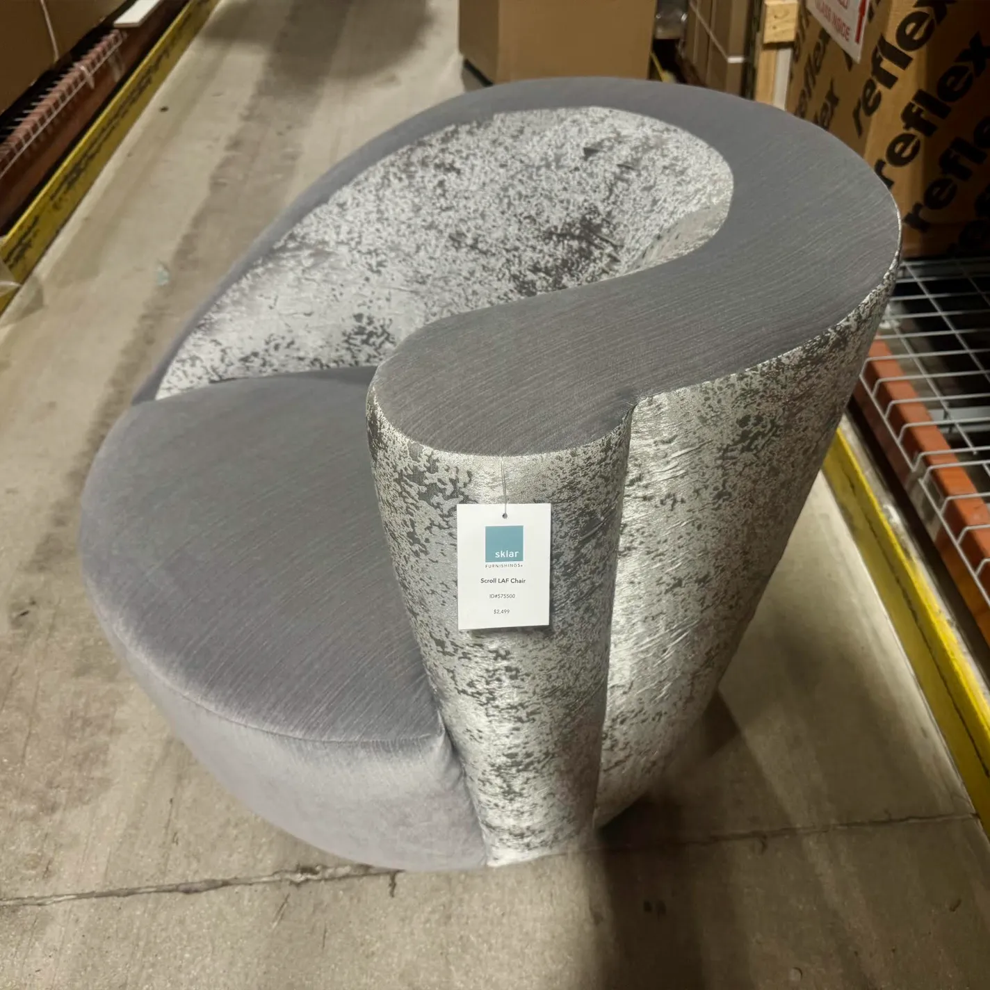 A modern, sculptural gray velvet chair with a silver textured back and armrest, featuring a scroll-like design, sits on a concrete floor in a warehouse.