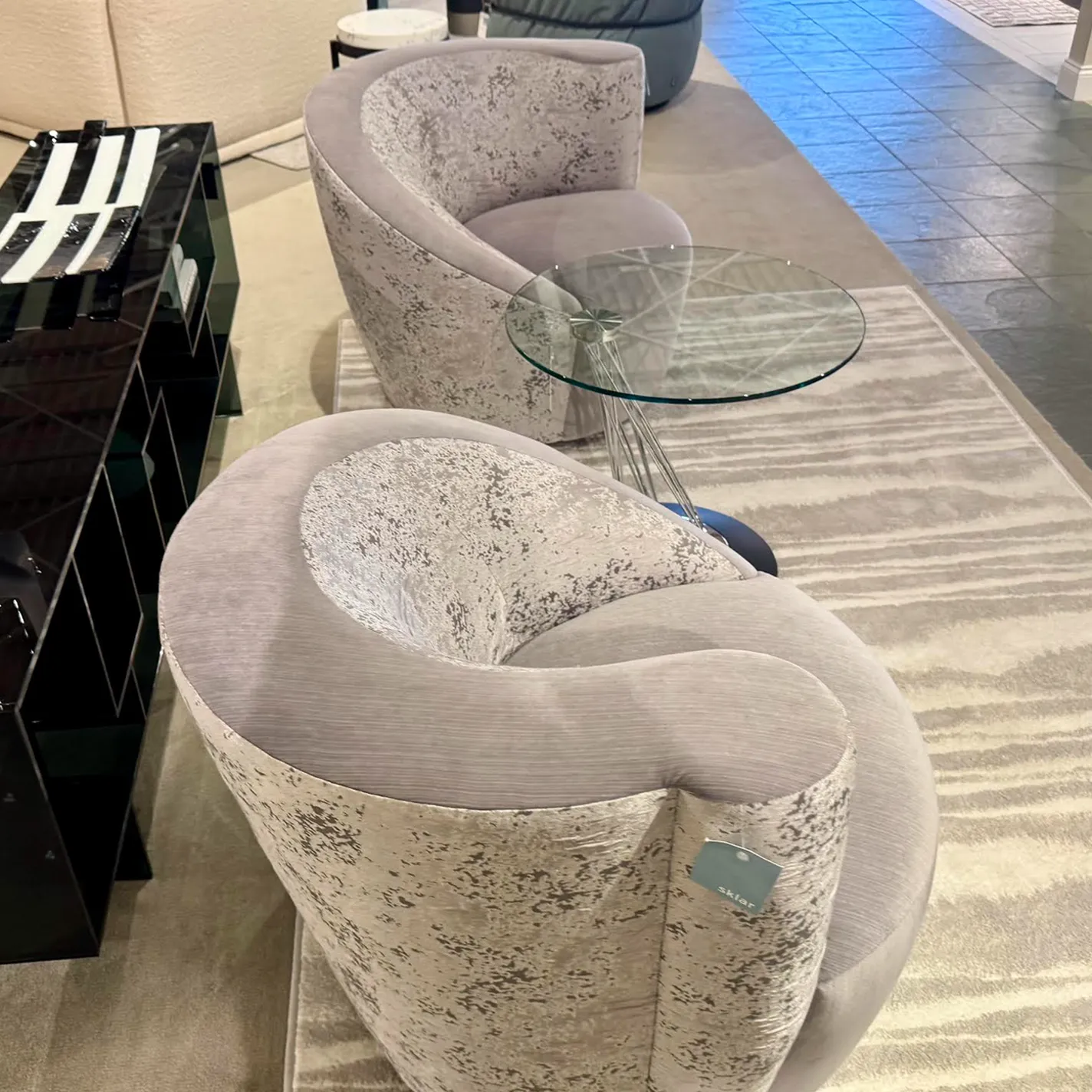 Two light gray and silver curved armchairs with a glass-top table between them, on a striped rug. A black console is to the left.