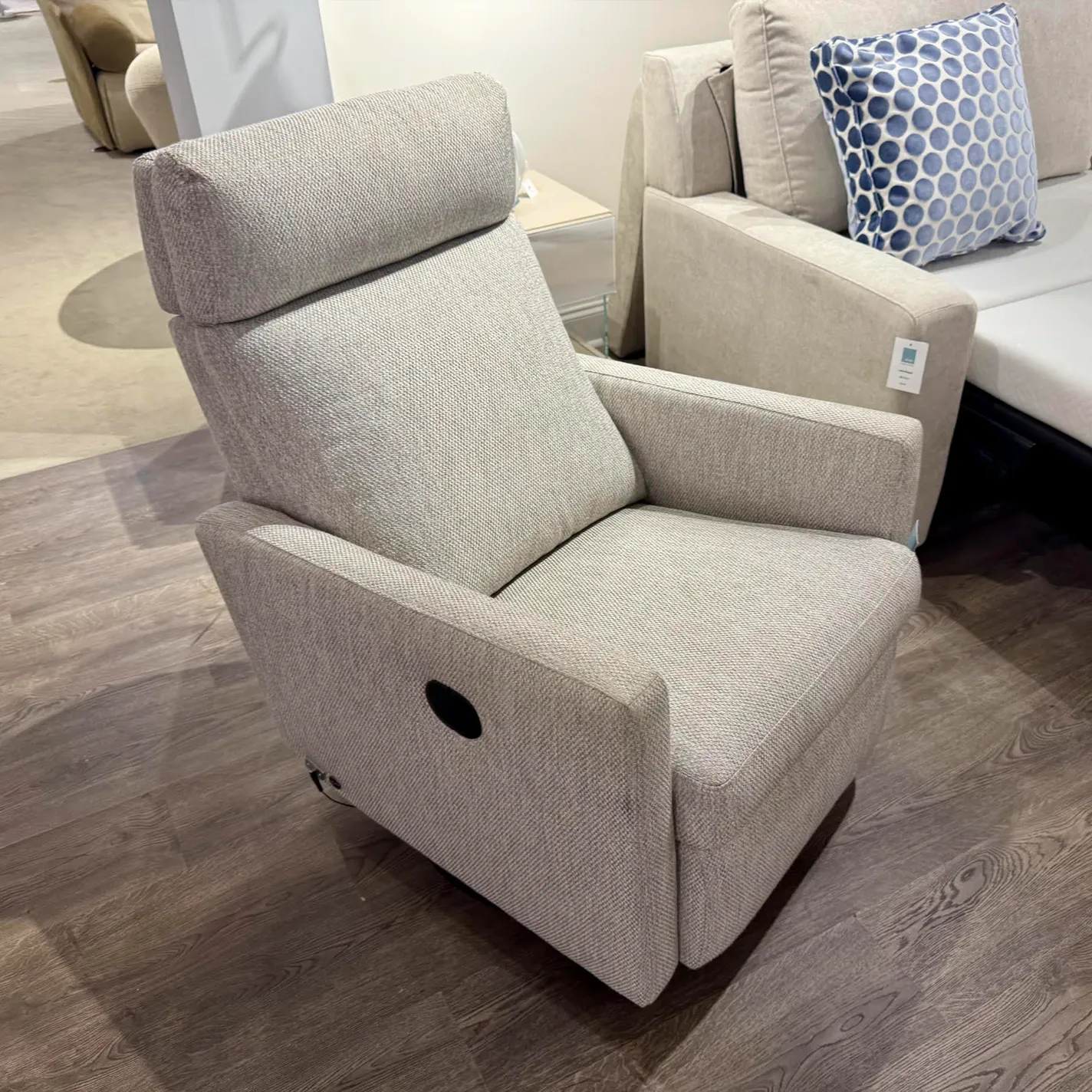 A light grey fabric recliner chair with a high back and black handle on a dark wood-look floor, with a beige sofa in the background.