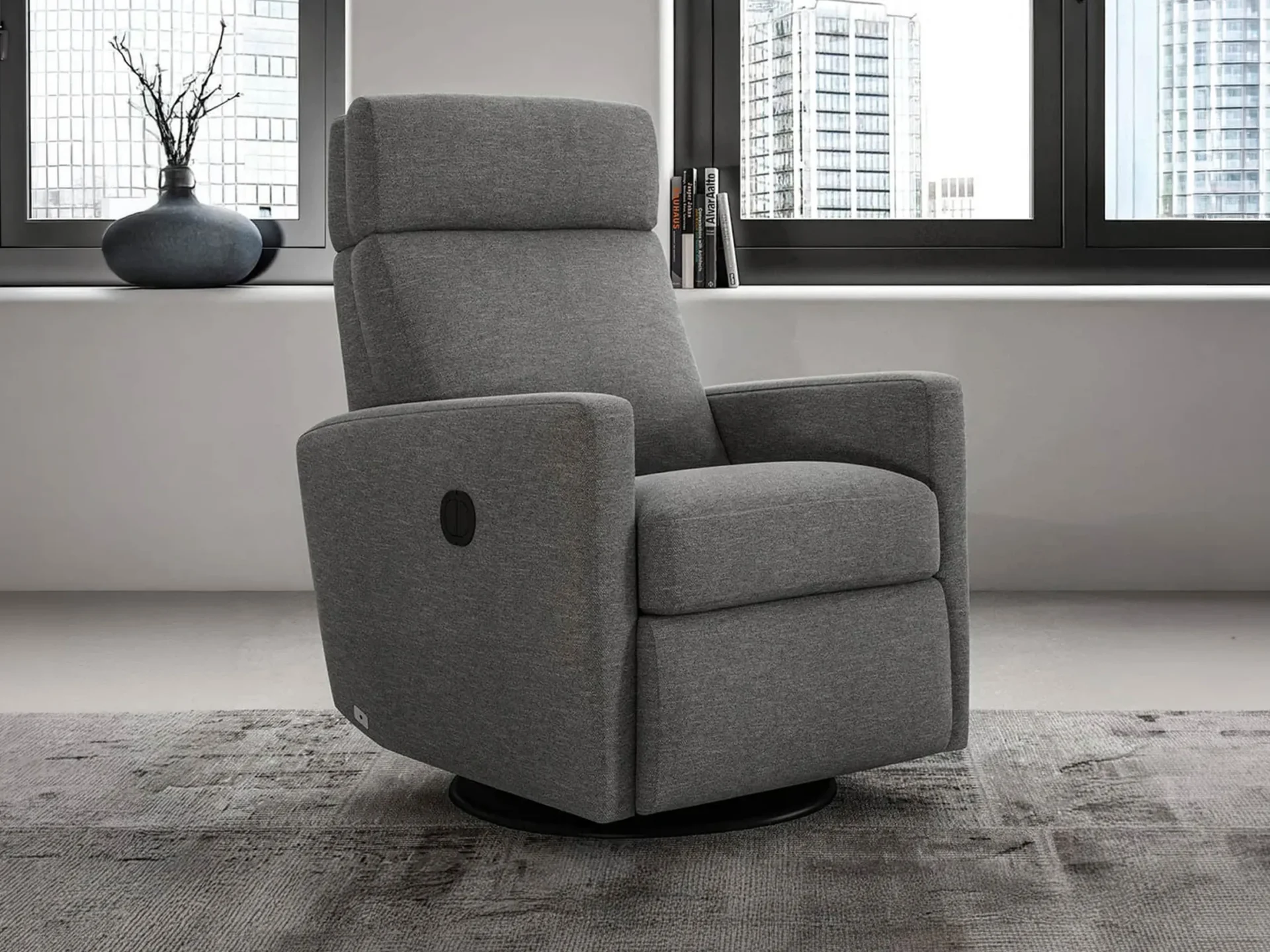 A gray fabric recliner with a black control panel and swivel base, positioned in front of a large window overlooking a city skyline. A vase with branches and a stack of books are on the window sill.