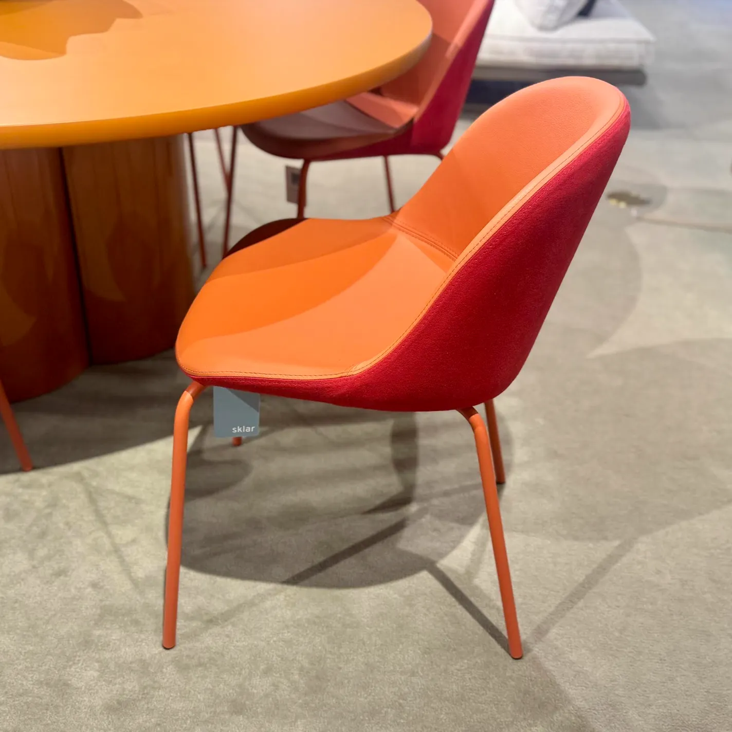A modern dining chair with an orange seat and red back, on orange metal legs, next to a matching glossy orange table. A 'sklar' tag hangs from the chair.