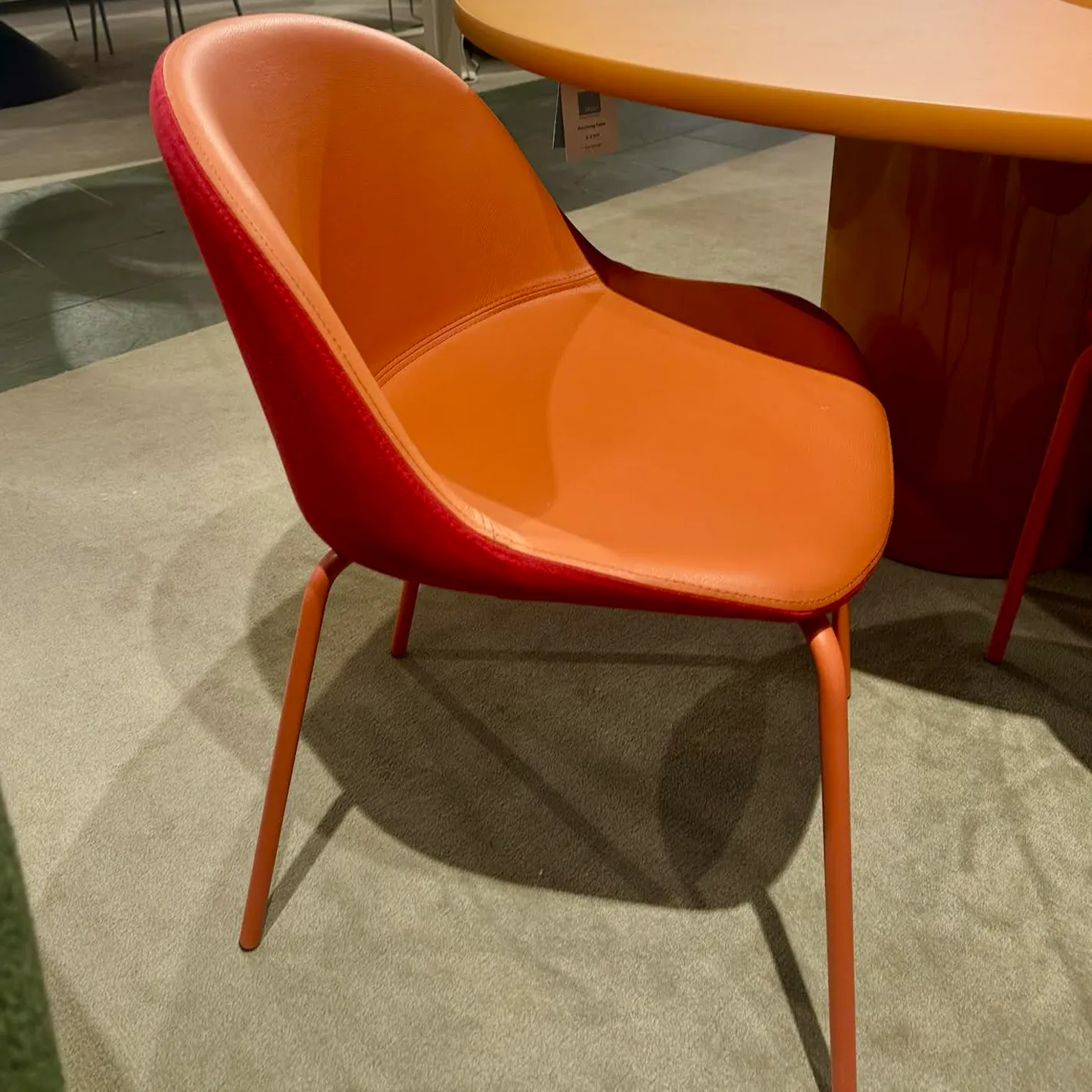 A modern chair with a bright orange seat and a reddish-orange fabric back, on slender orange metal legs. It sits on a light gray carpet next to a matching round orange table.