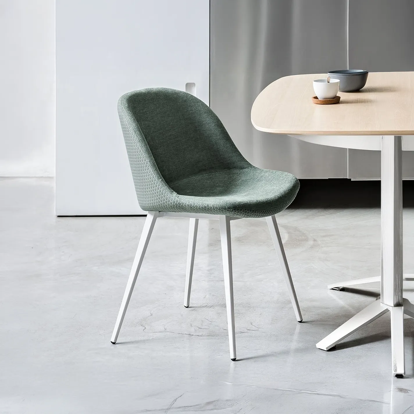 A modern sage green upholstered dining chair with white metal legs next to a light wood table in a minimalist room.