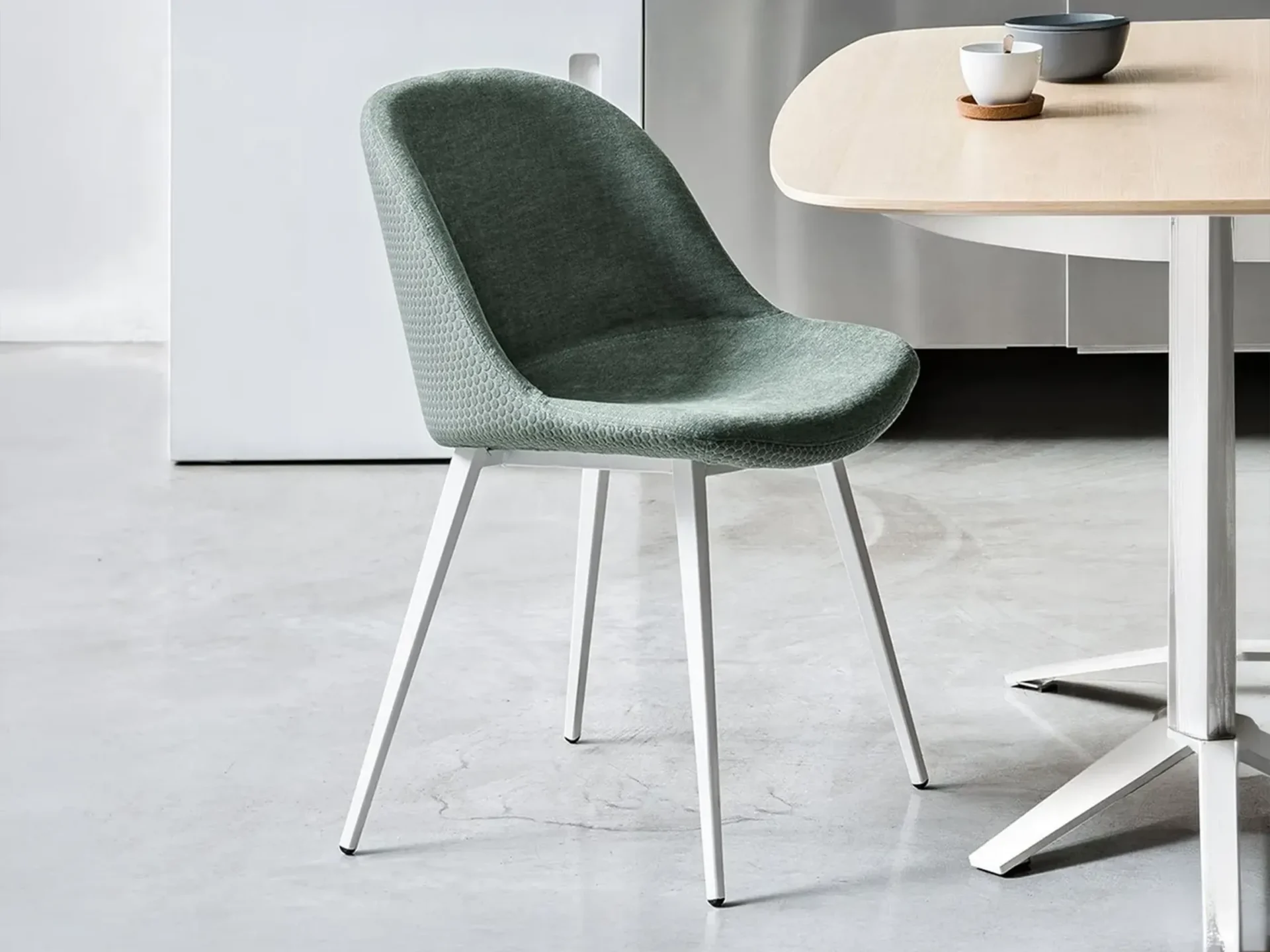 A modern sage green upholstered dining chair with white metal legs next to a light wood table with a cup and bowl in a minimalist room.