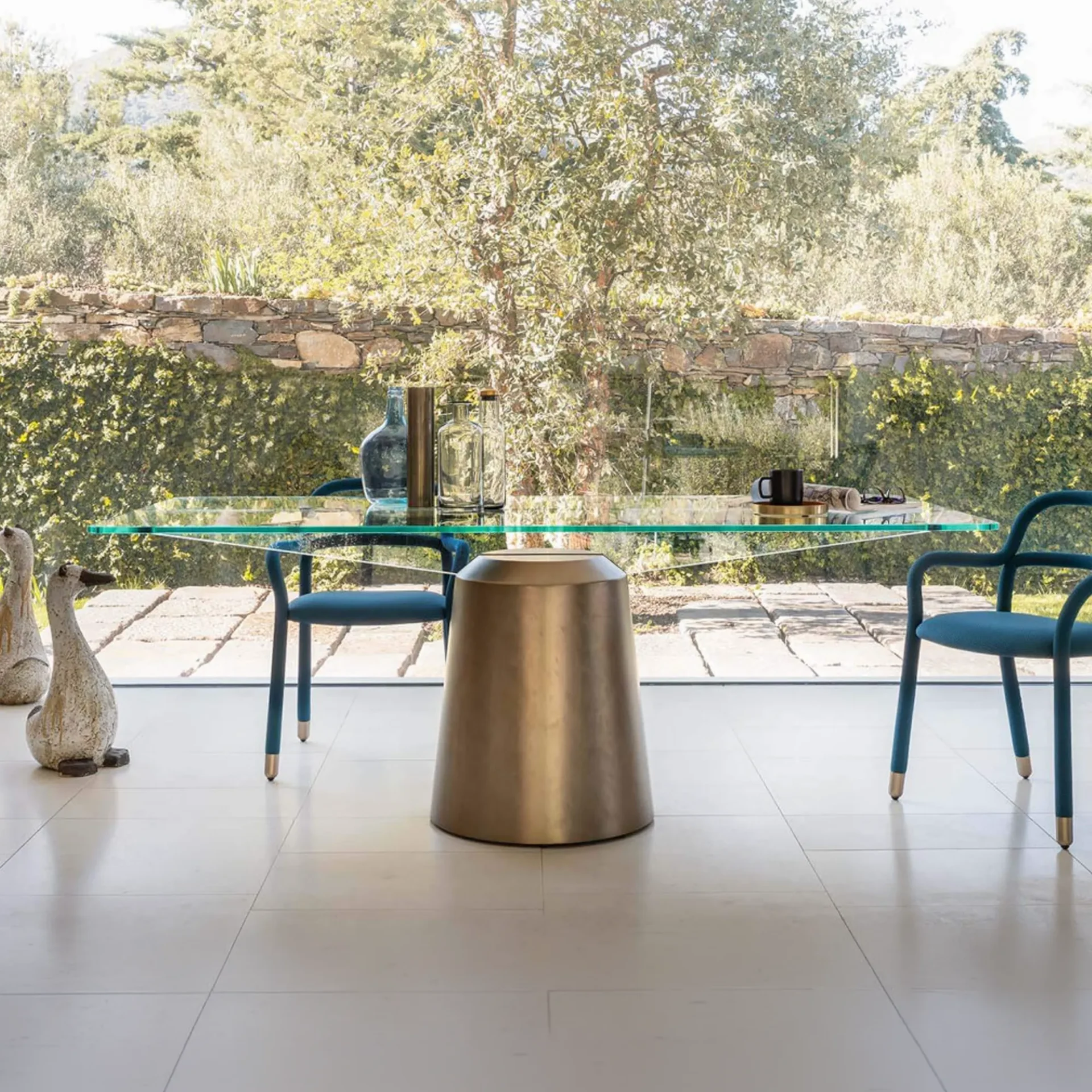 A modern dining room with a glass table, brass base, and teal chairs, overlooking a green garden through a large window.