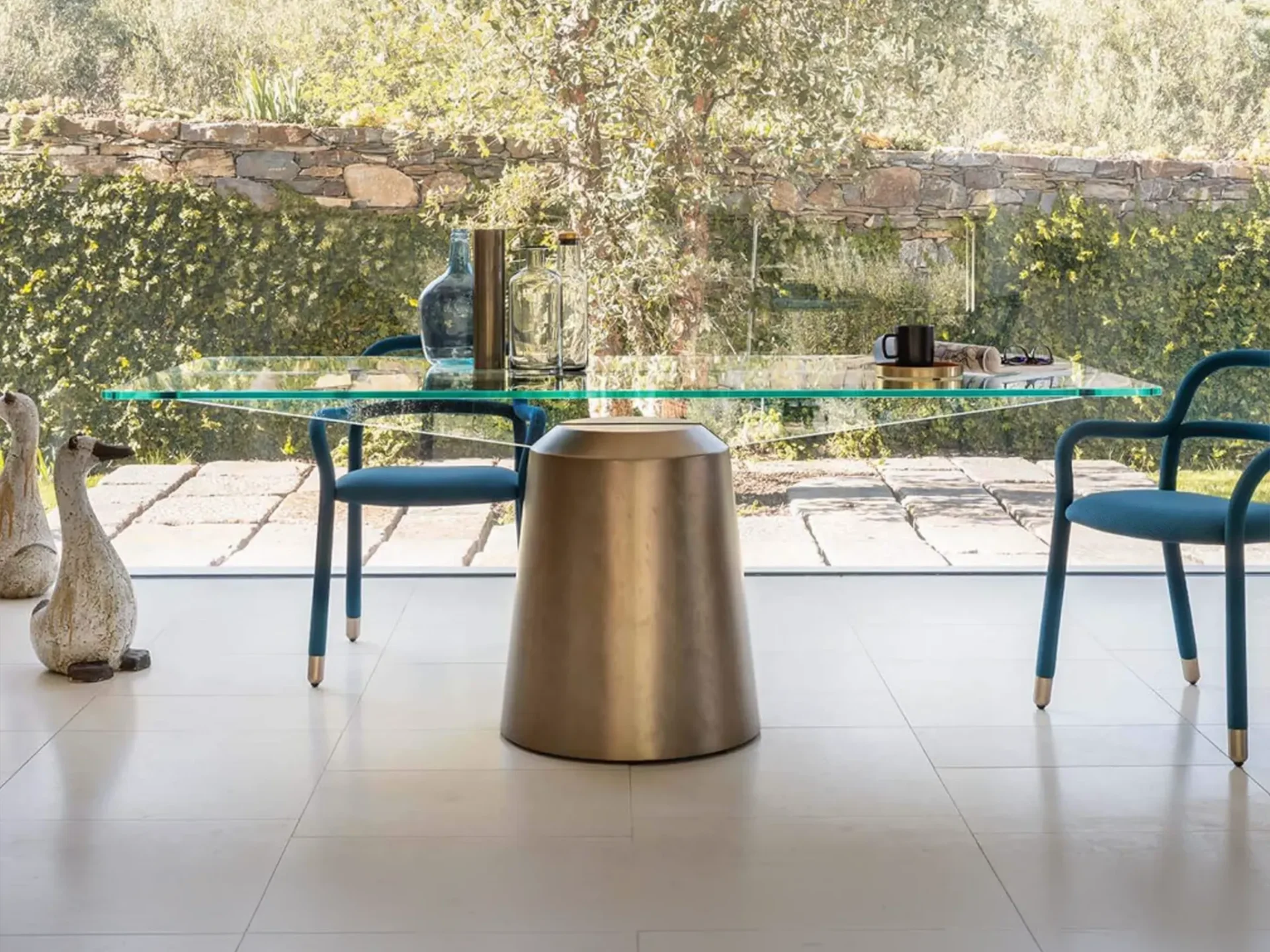 A modern dining table with a glass top and bronze base, flanked by two teal chairs, in front of a large window overlooking a lush green garden.