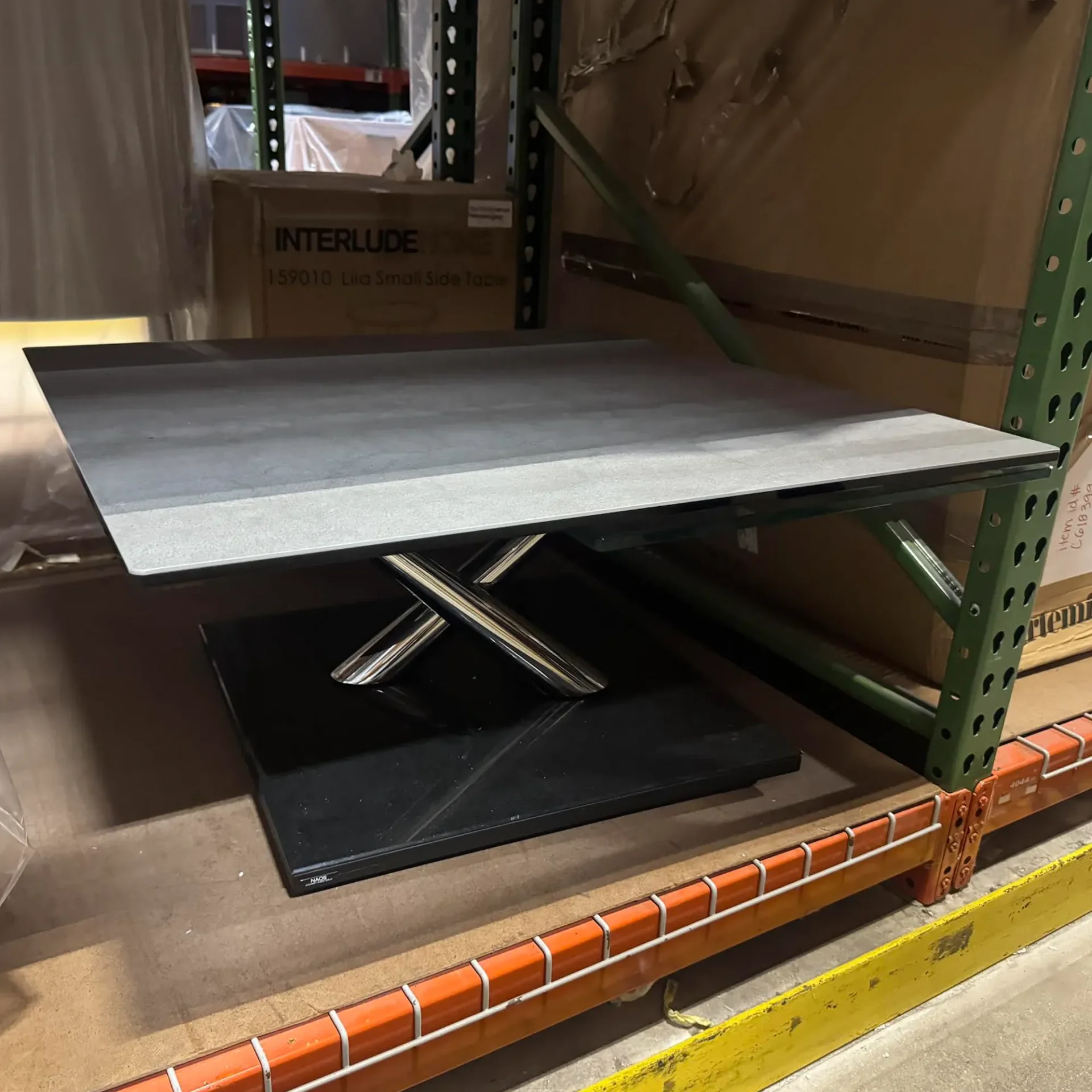 A modern dark gray rectangular side table with a chrome X-shaped base sits on a warehouse shelf next to its cardboard box packaging.