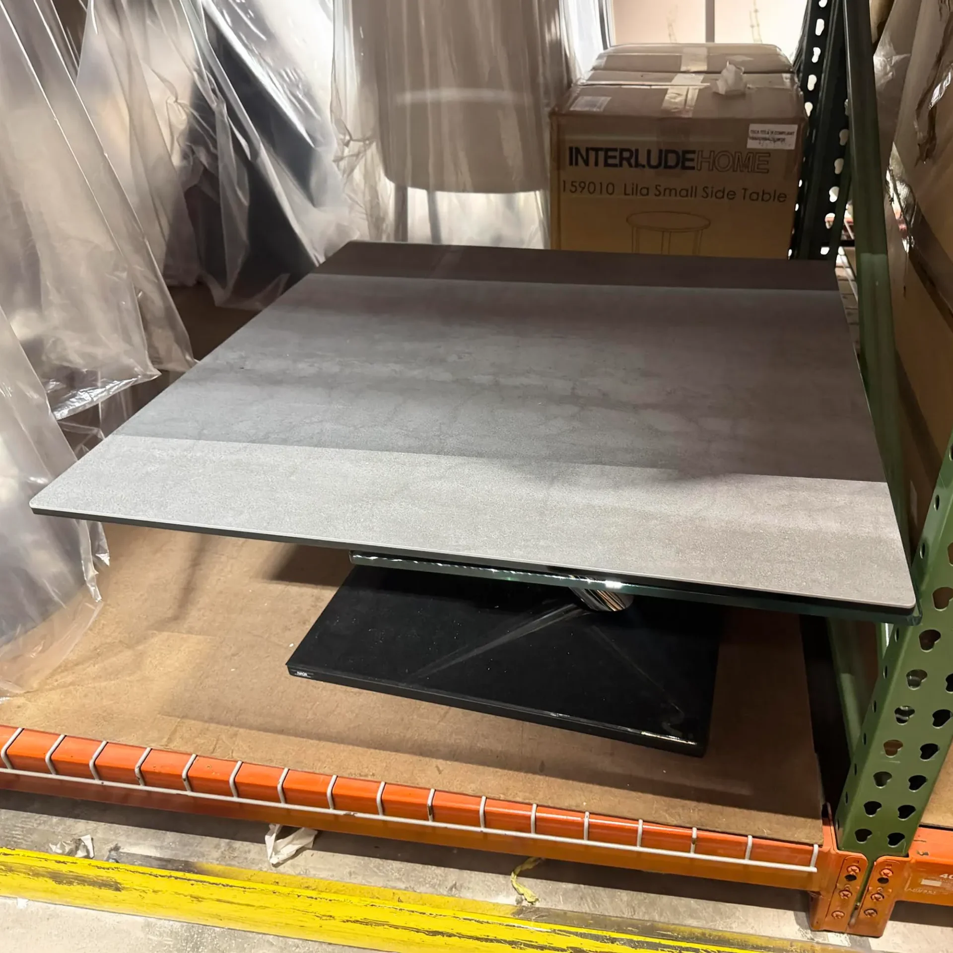 A gray rectangular table with a glass and black base sits on a brown shelf in a warehouse. A box labeled 'InterludeHome Lila Small Side Table' is visible in the background.