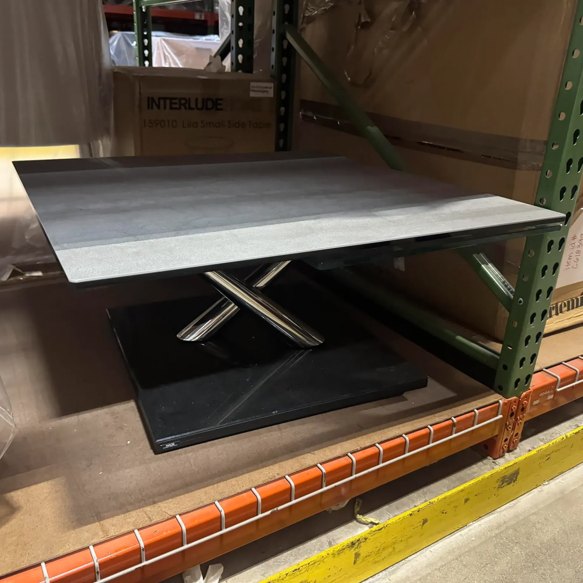 A modern side table with a gray top, chrome X-shaped legs, and a black base, sitting on a concrete floor in a warehouse setting with shelving and boxes in the background.