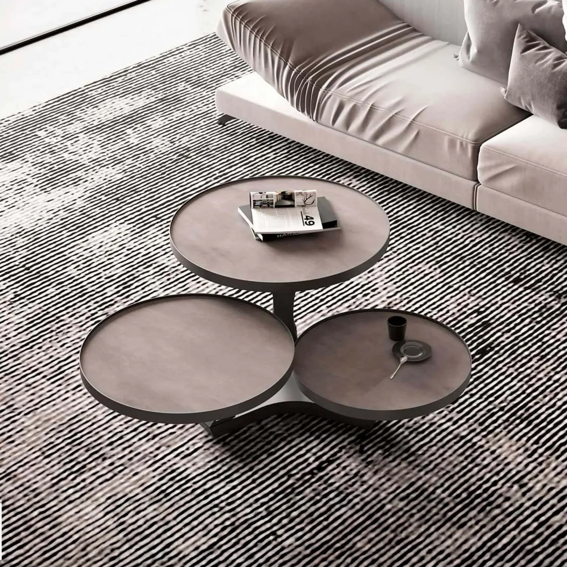 A modern three-tiered circular coffee table with books and a cup, set on a black and white striped rug next to a light-colored sofa.