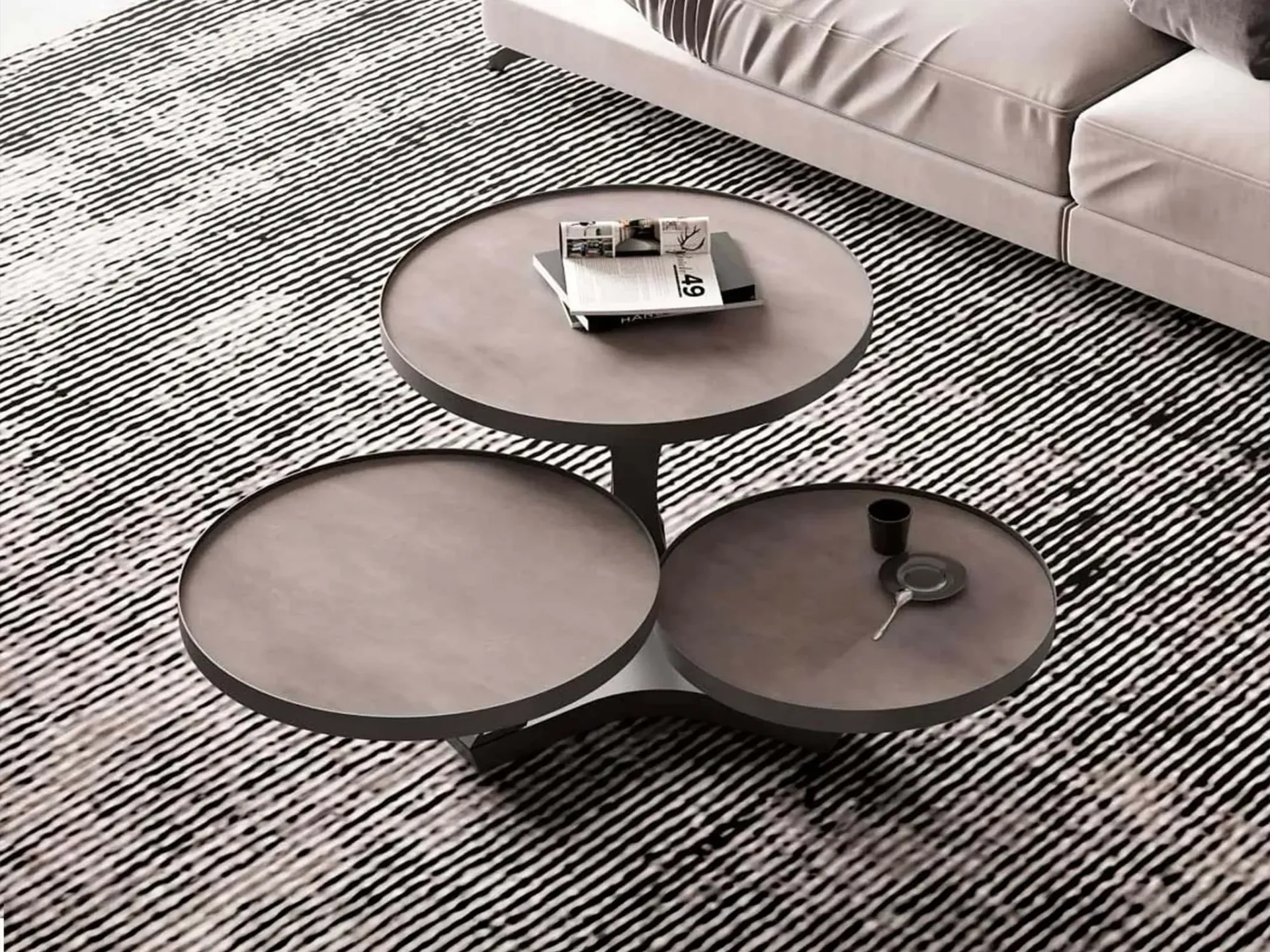 A modern, three-tiered coffee table with circular grey-brown tops on a black and white striped rug. Books are on the top tier, and a cup and saucer are on a lower tier. A light-colored sofa is in the background.