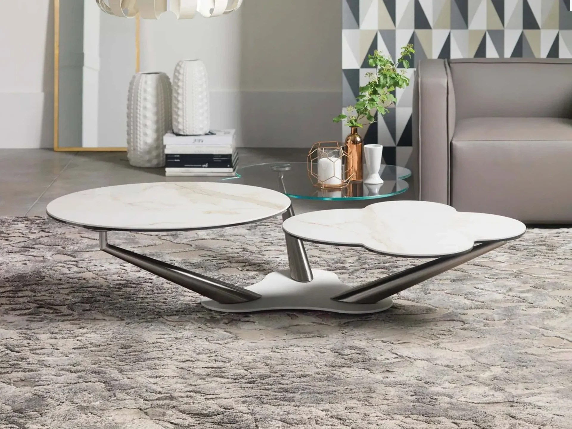 A modern living room with a two-tiered coffee table featuring white marble-patterned tops and a metal base, on a gray textured rug. A gray sofa and geometric wallpaper are in the background.