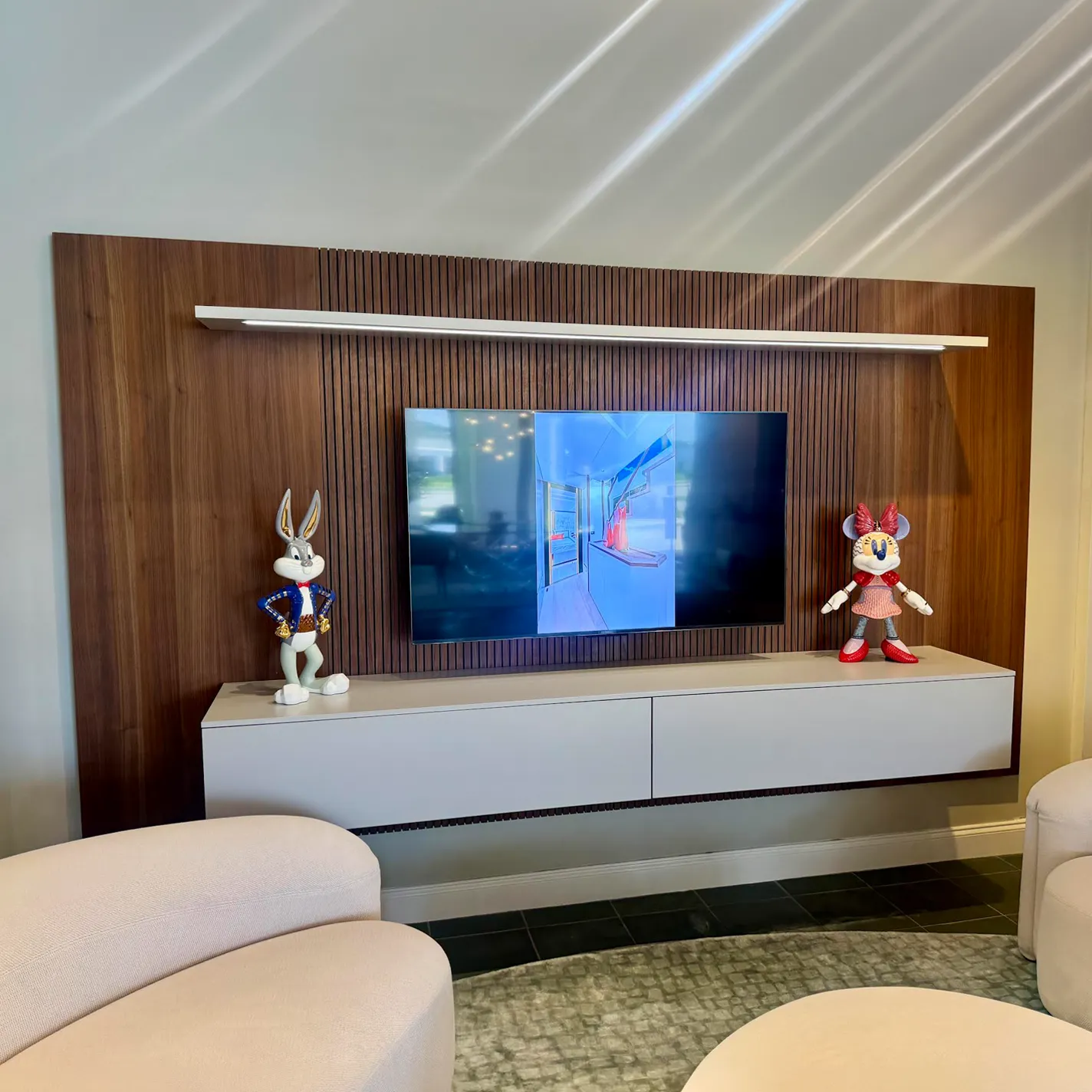 A modern living room with a TV mounted on a wood-paneled wall, a floating console, and Bugs Bunny and Minnie Mouse figurines.