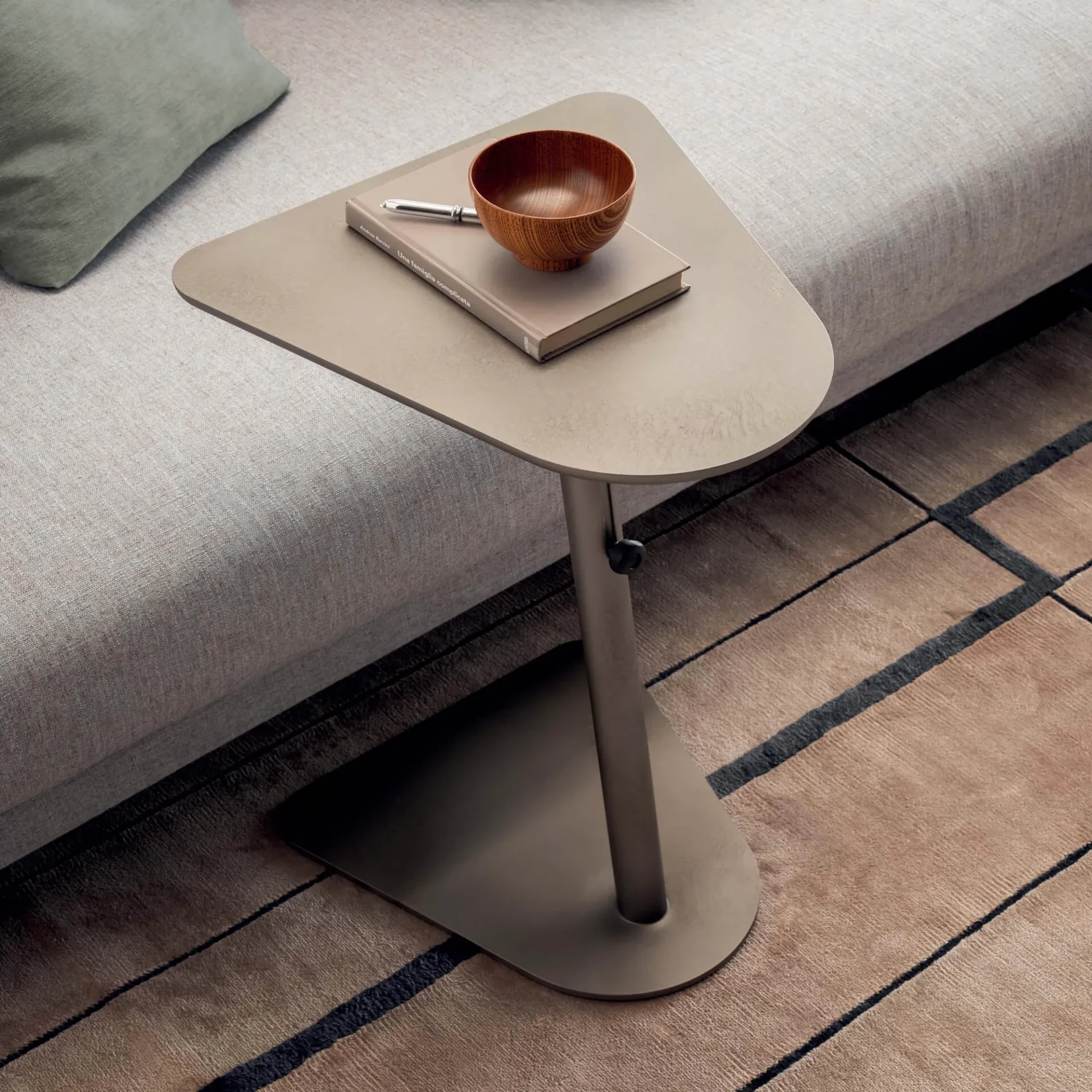 A modern taupe C-shaped side table next to a light beige sofa. On the table are a book, a silver pen, and a wooden bowl. A geometric rug is on the floor.